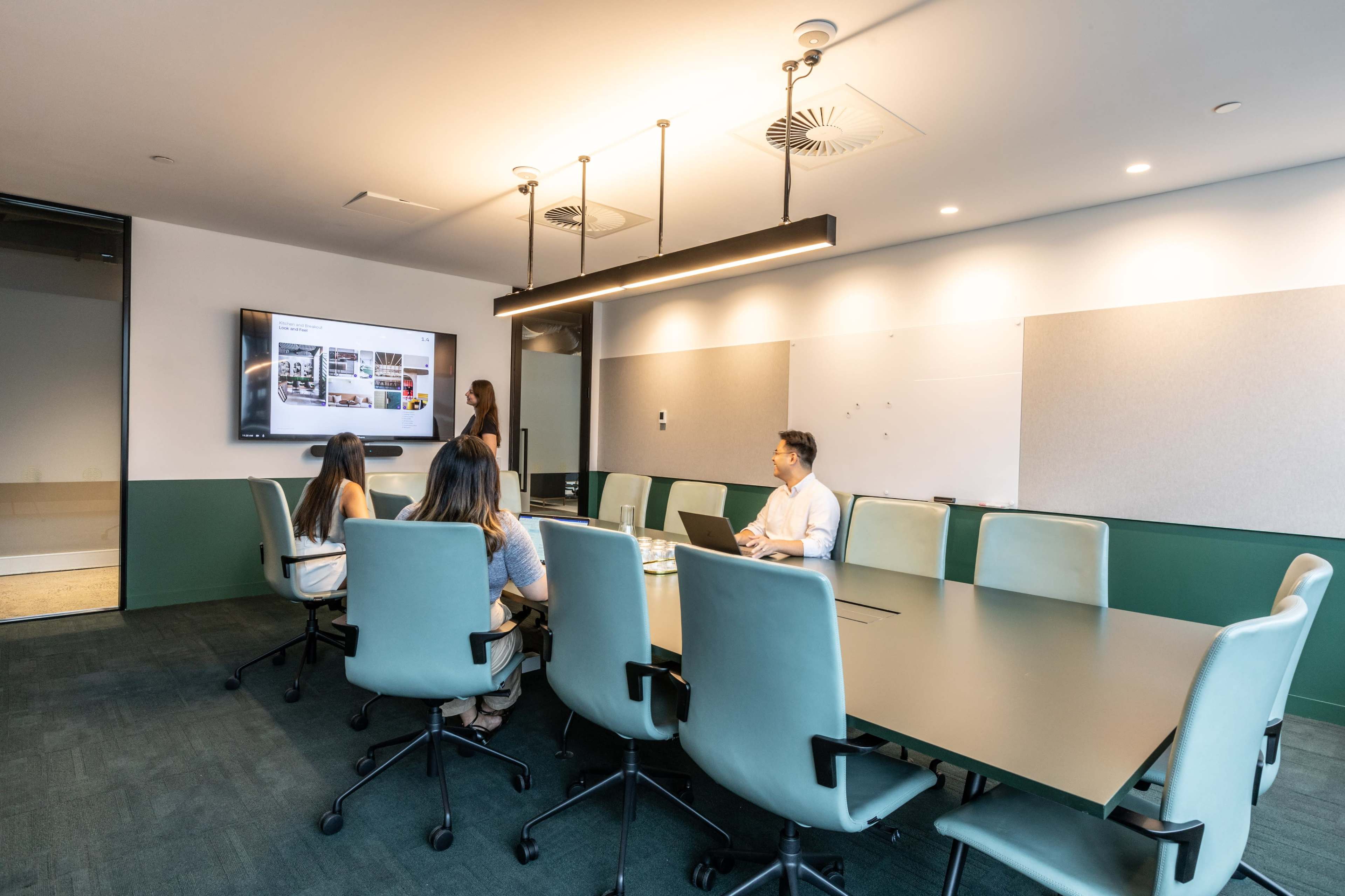 A group of five people sits around a large conference table in a modern meeting room, with a screen displaying a presentation.