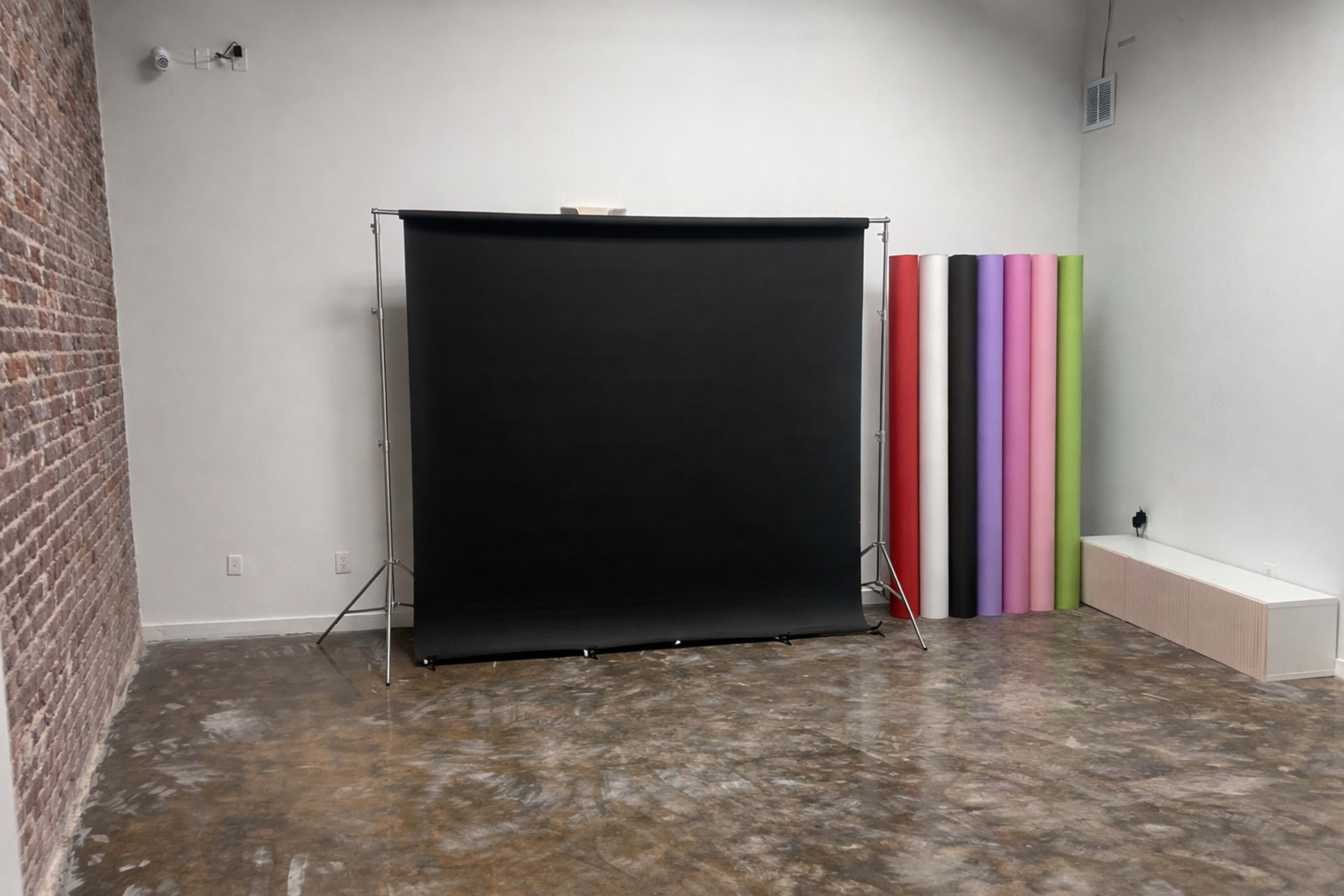 The image shows a photography studio with a black backdrop setup on a stand and a vertical array of colorful paper rolls in various shades alongside a wall.