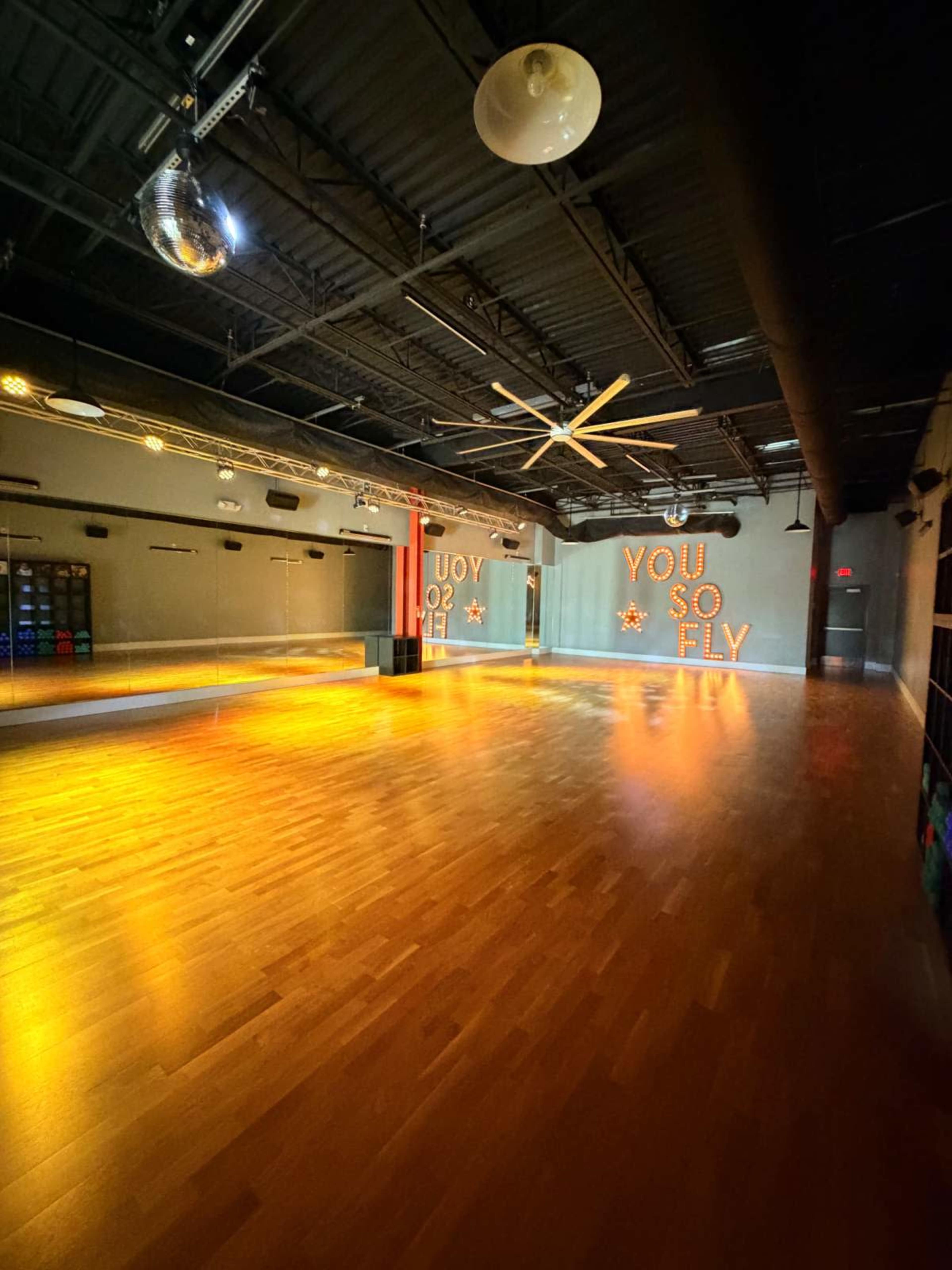 The image shows an empty dance studio with wooden flooring, mirrored walls, and the words "YOU SO FLY" illuminated on one wall.