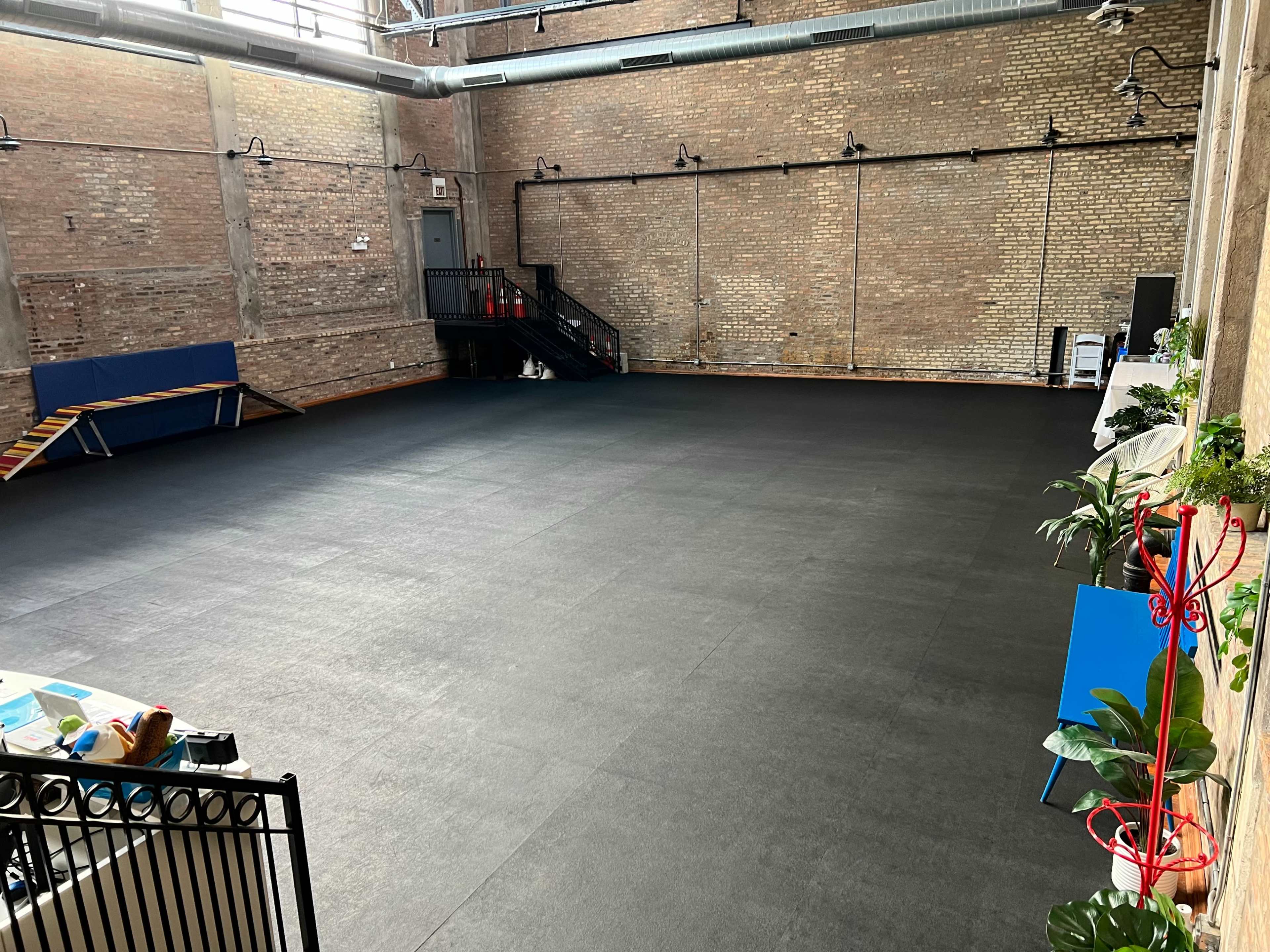 A spacious interior room with brick walls and a black rubber floor, featuring a staircase on one side and some furniture, including potted plants, on another side.