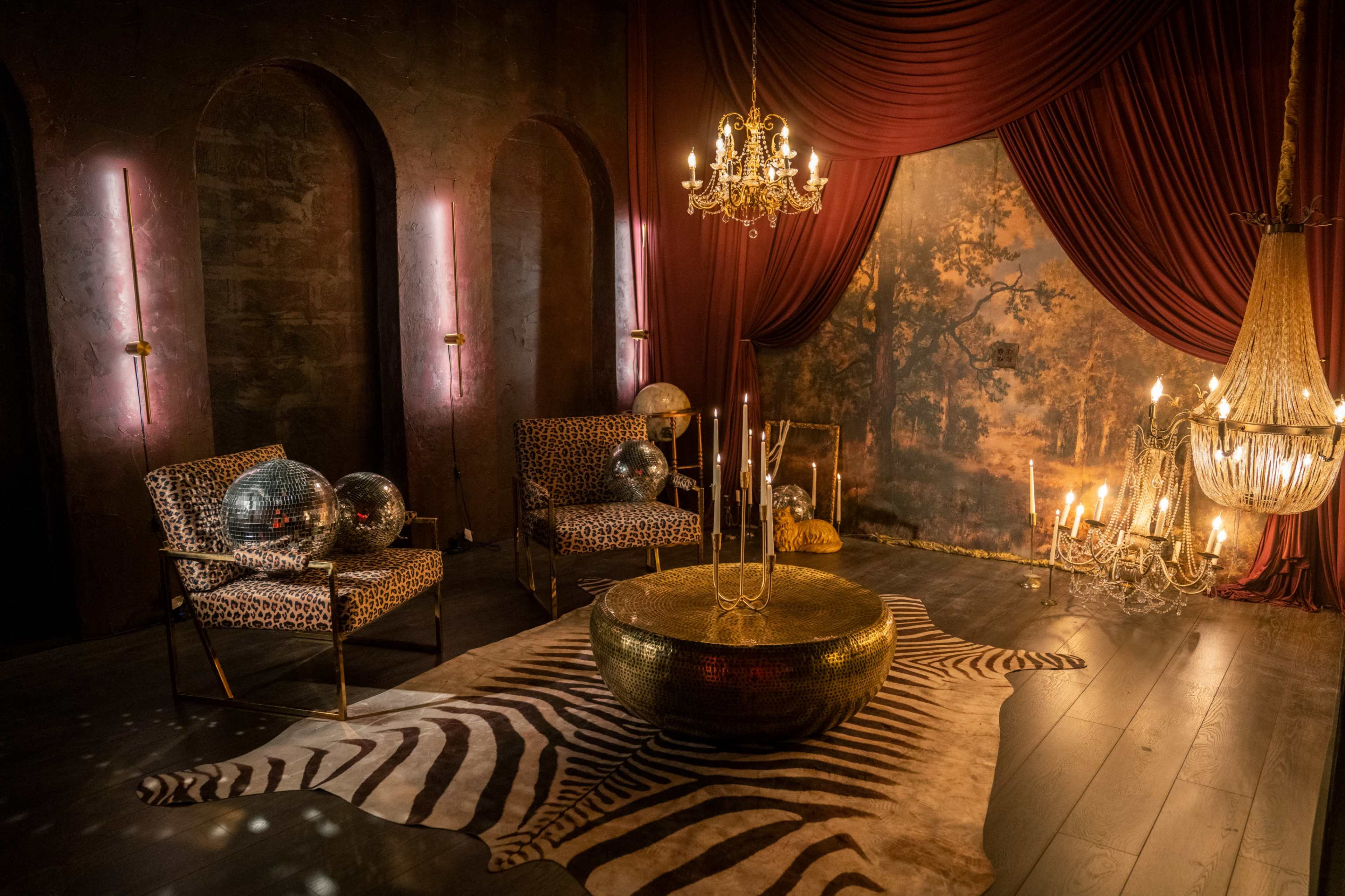 The scene features a stylish interior with animal print chairs, a gold circular coffee table on a zebra-striped rug, and soft lighting from candles and a chandelier, set against a backdrop of draped fabric and a painted mural.