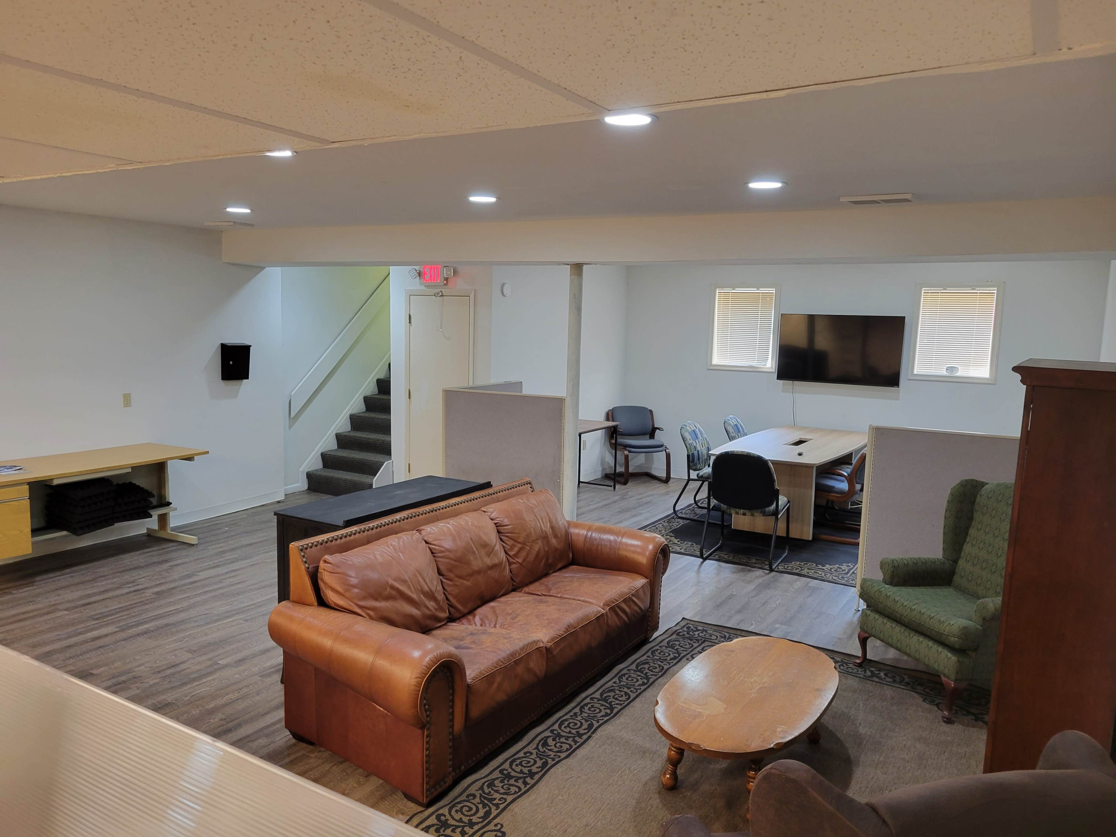 A spacious, multi-purpose room with a brown leather sofa, a wooden coffee table, and various seating arrangements, including a desk and an area with a television.