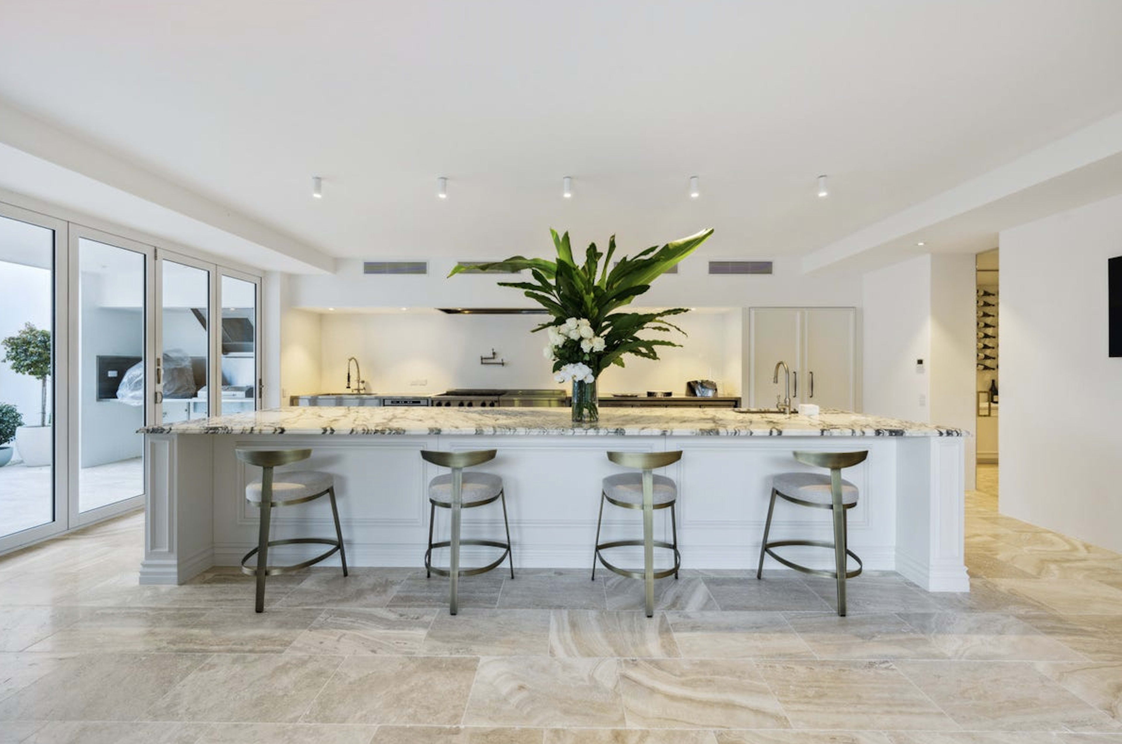 A modern kitchen features a large island with a marble countertop and bar stools, surrounded by large windows that let in natural light.