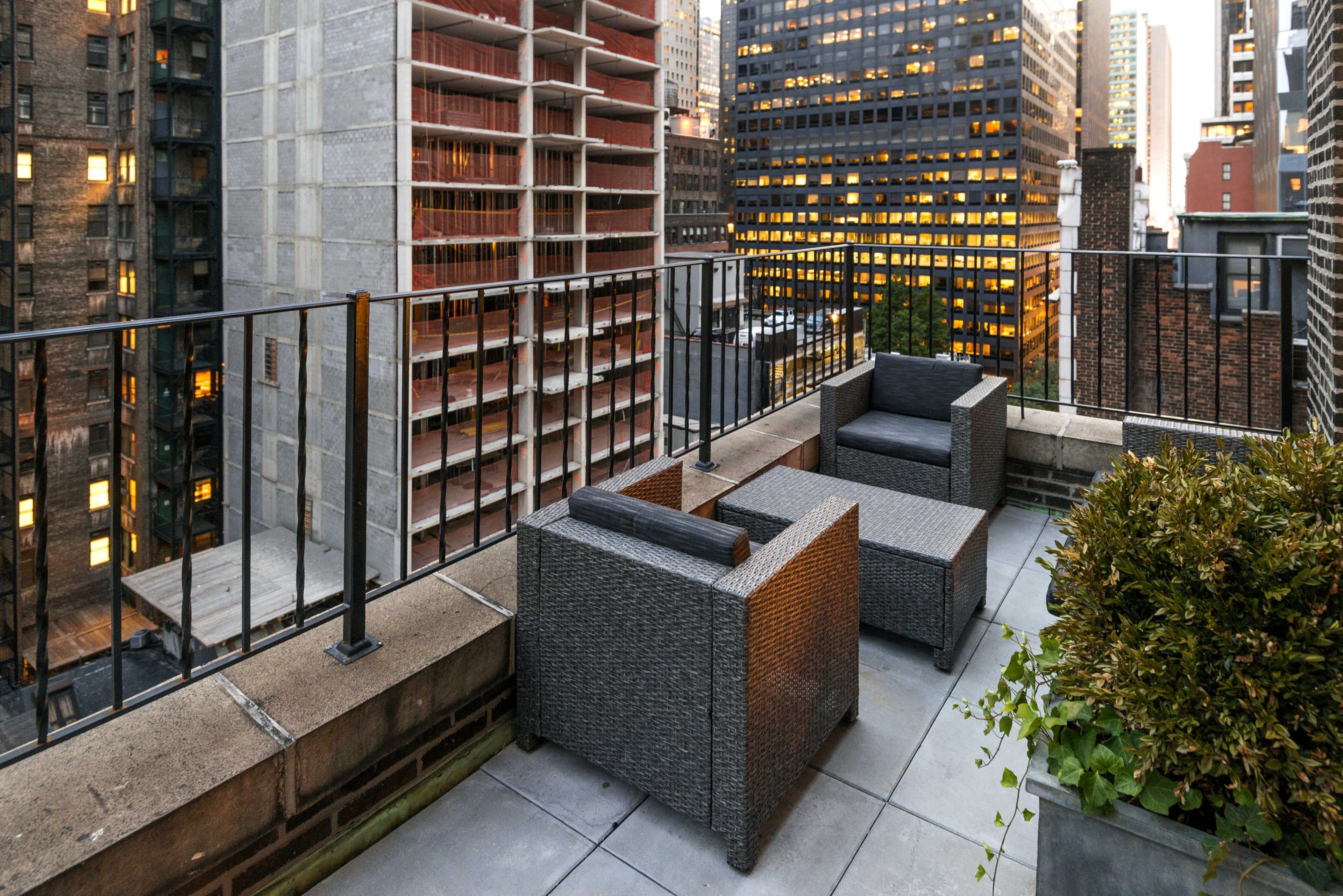 The image shows a rooftop terrace with two wicker chairs overlooking a city skyline during twilight.