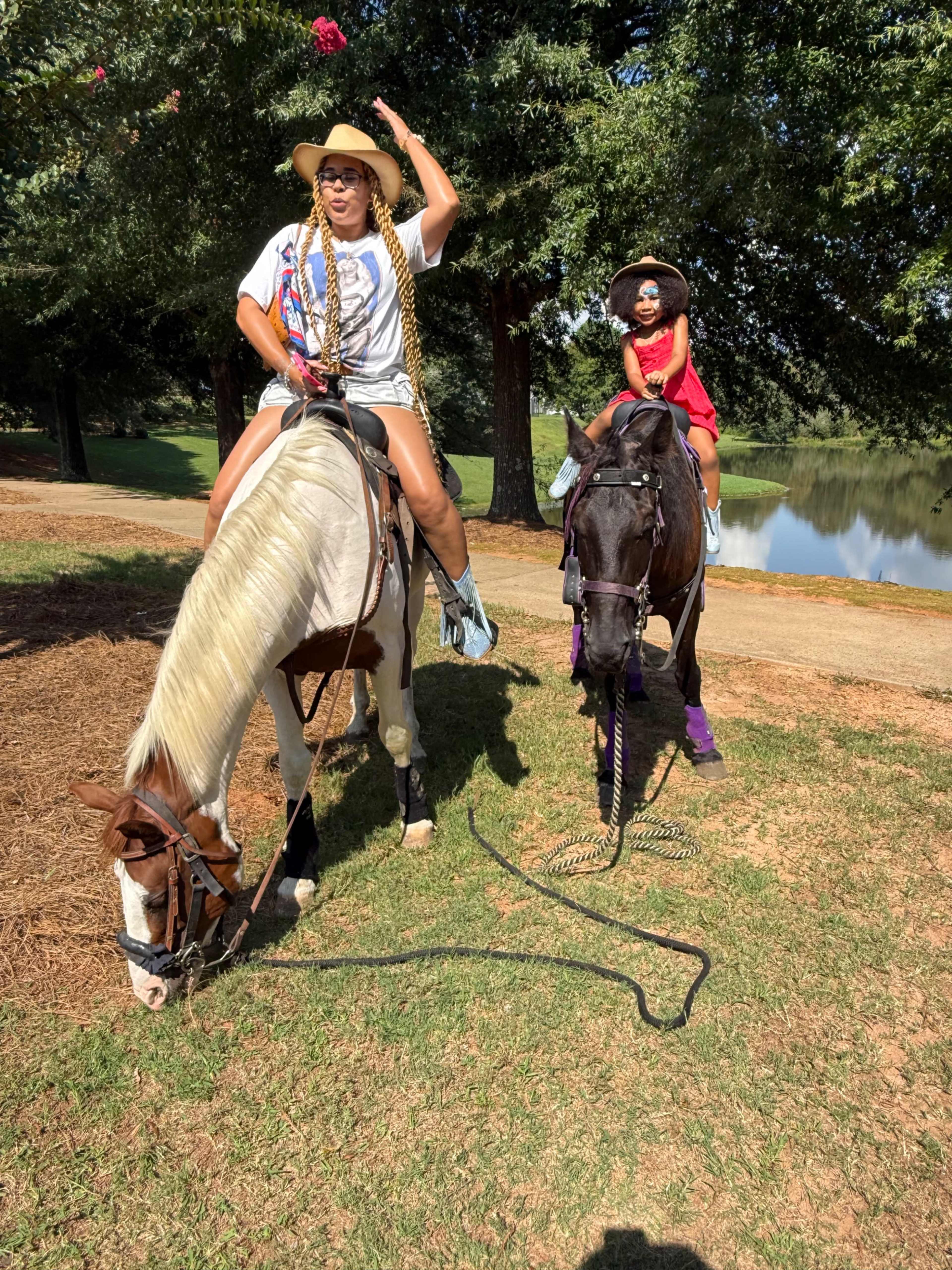 Two riders on horseback, one wearing a straw hat and sunglasses, and a child in a red outfit, are posed near a lake surrounded by trees.
