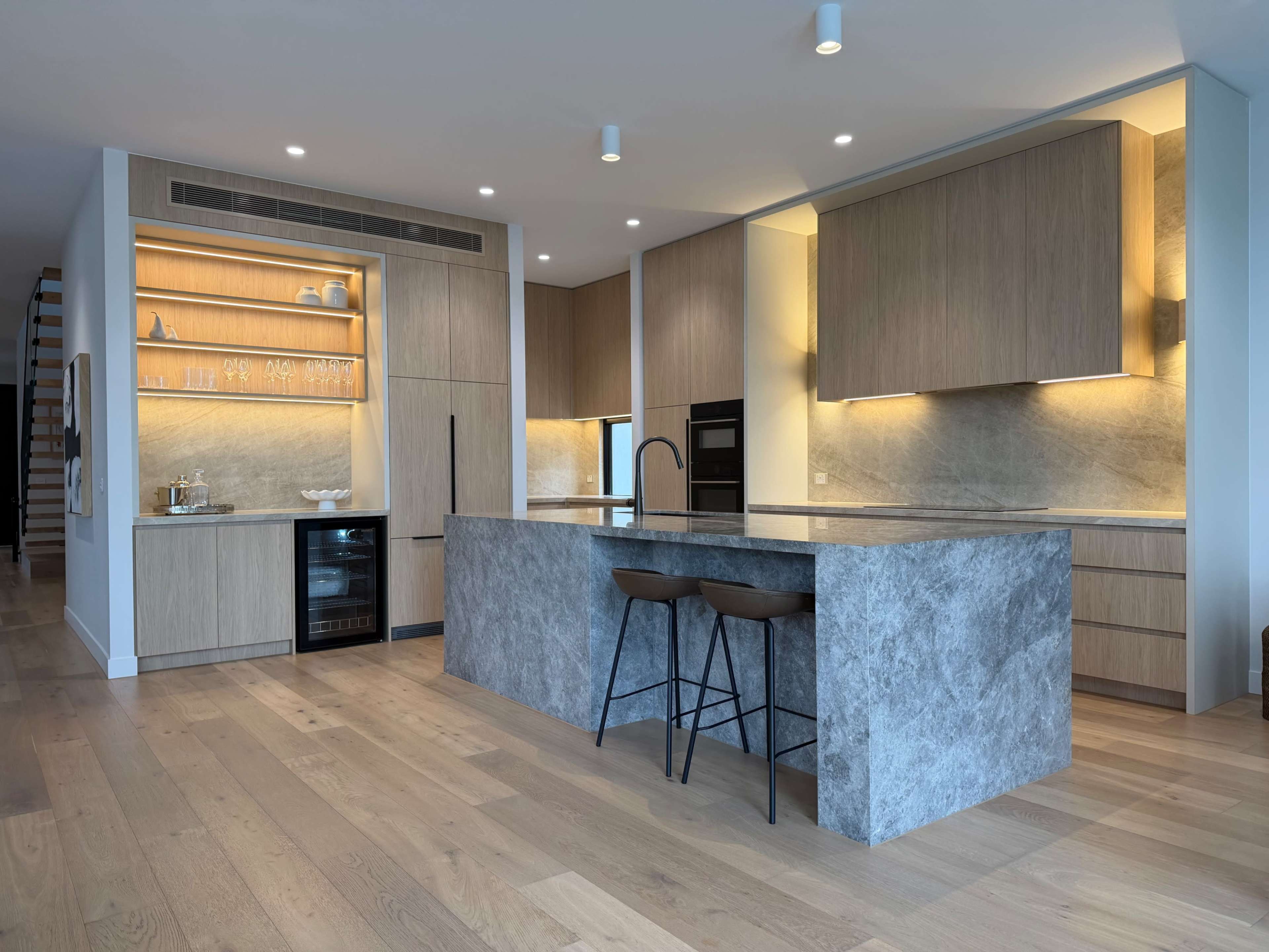 The image shows a modern kitchen featuring wood cabinetry, a stone countertop island with two bar stools, and warm ambient lighting.