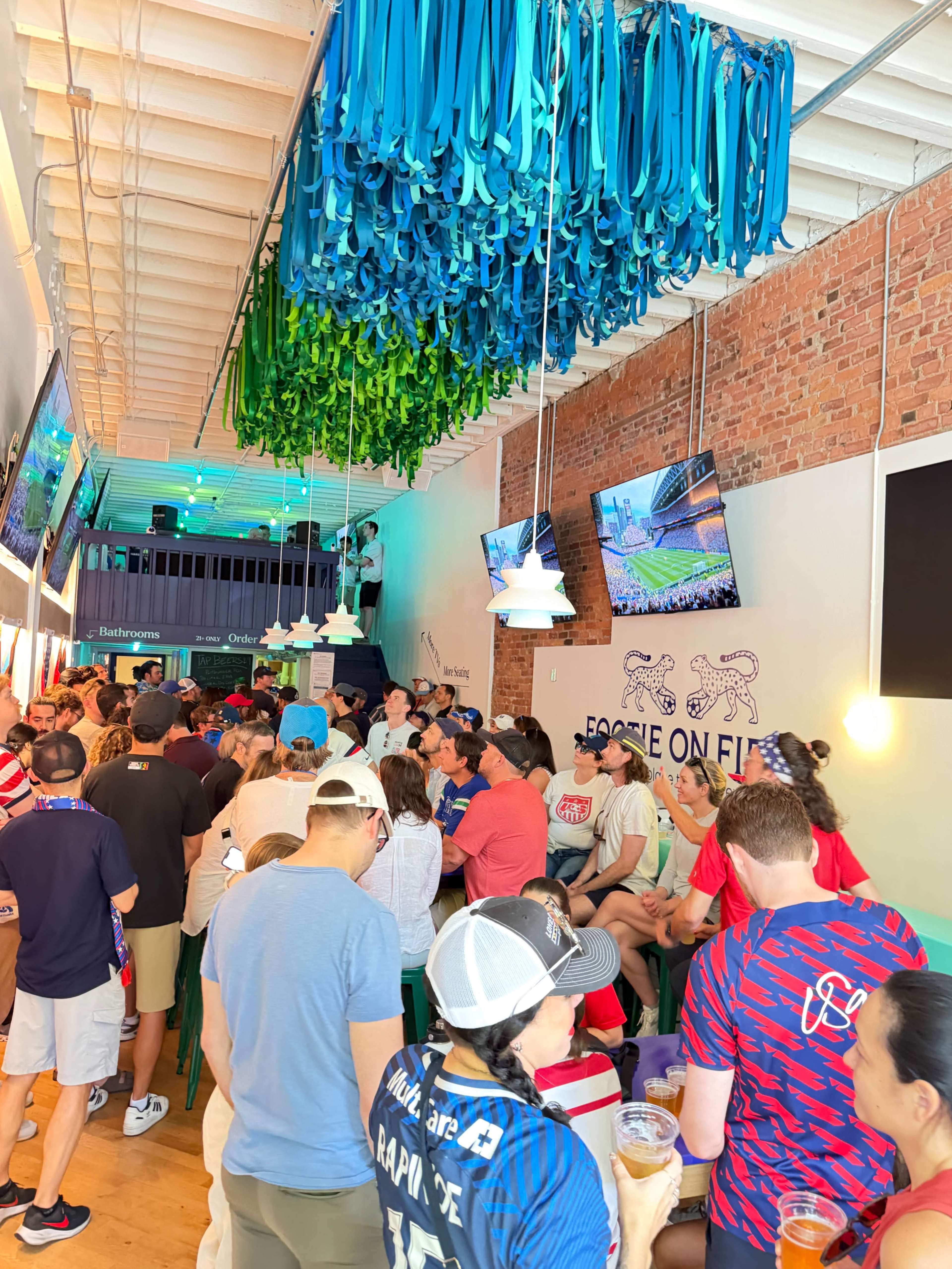 The image shows a crowded sports bar filled with patrons watching a game on multiple screens, adorned with vibrant decorations hanging from the ceiling.