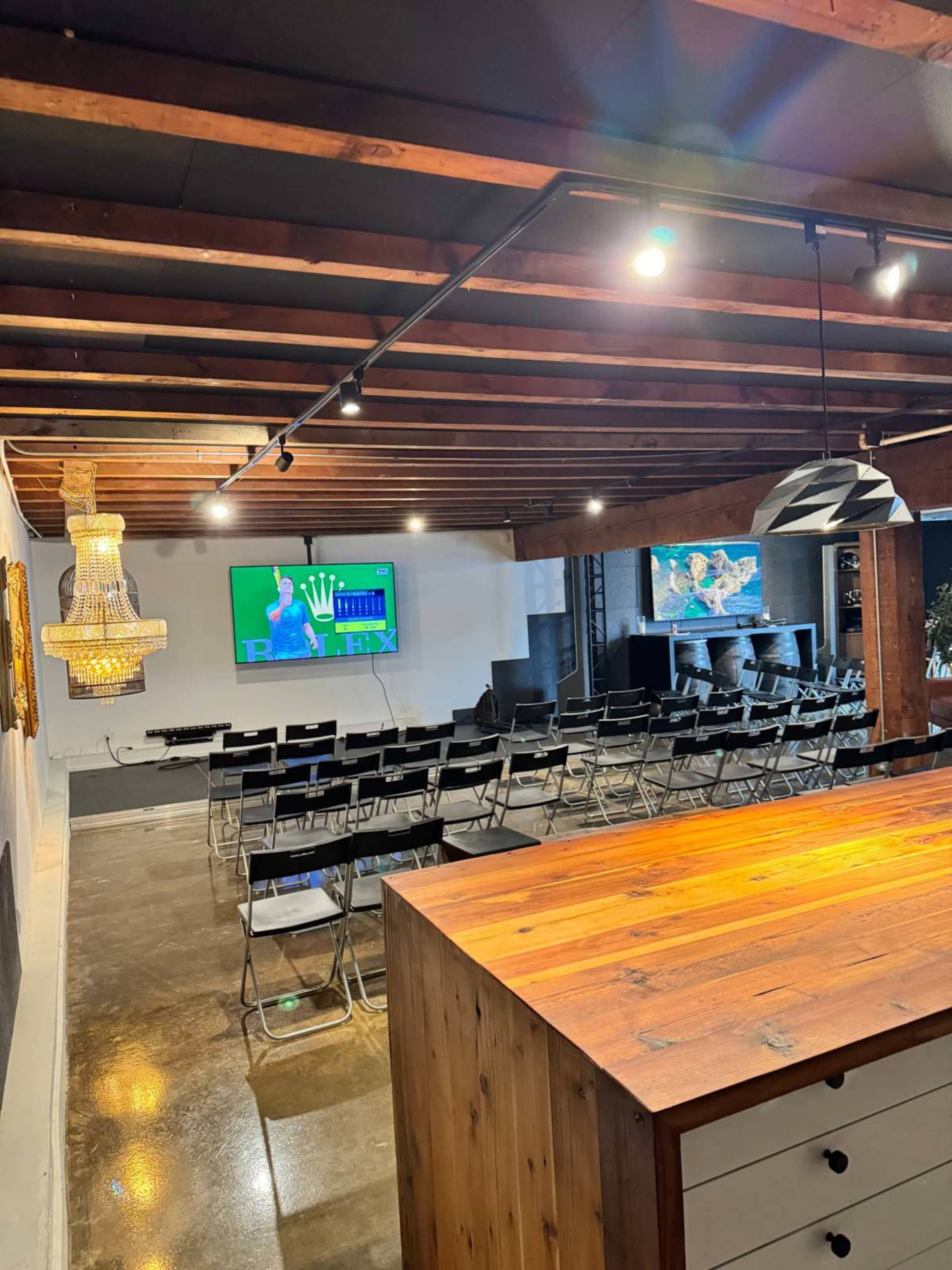 The image shows a modern conference room with rows of black chairs facing a television screen, exposed wooden beams, and a wooden counter.