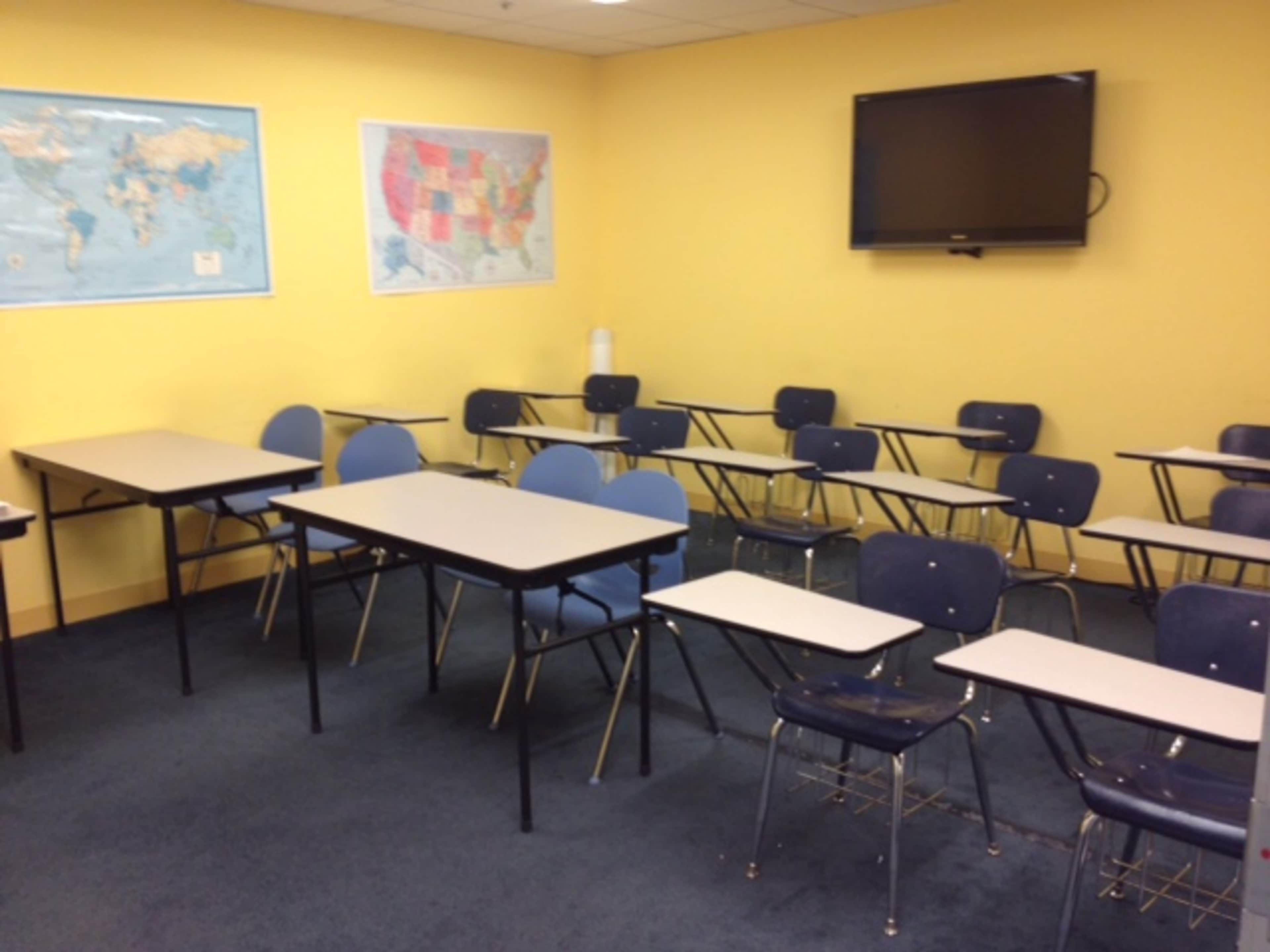 The image shows a classroom with several tables and chairs arranged in a semi-circle, featuring maps on the walls and a television mounted above.