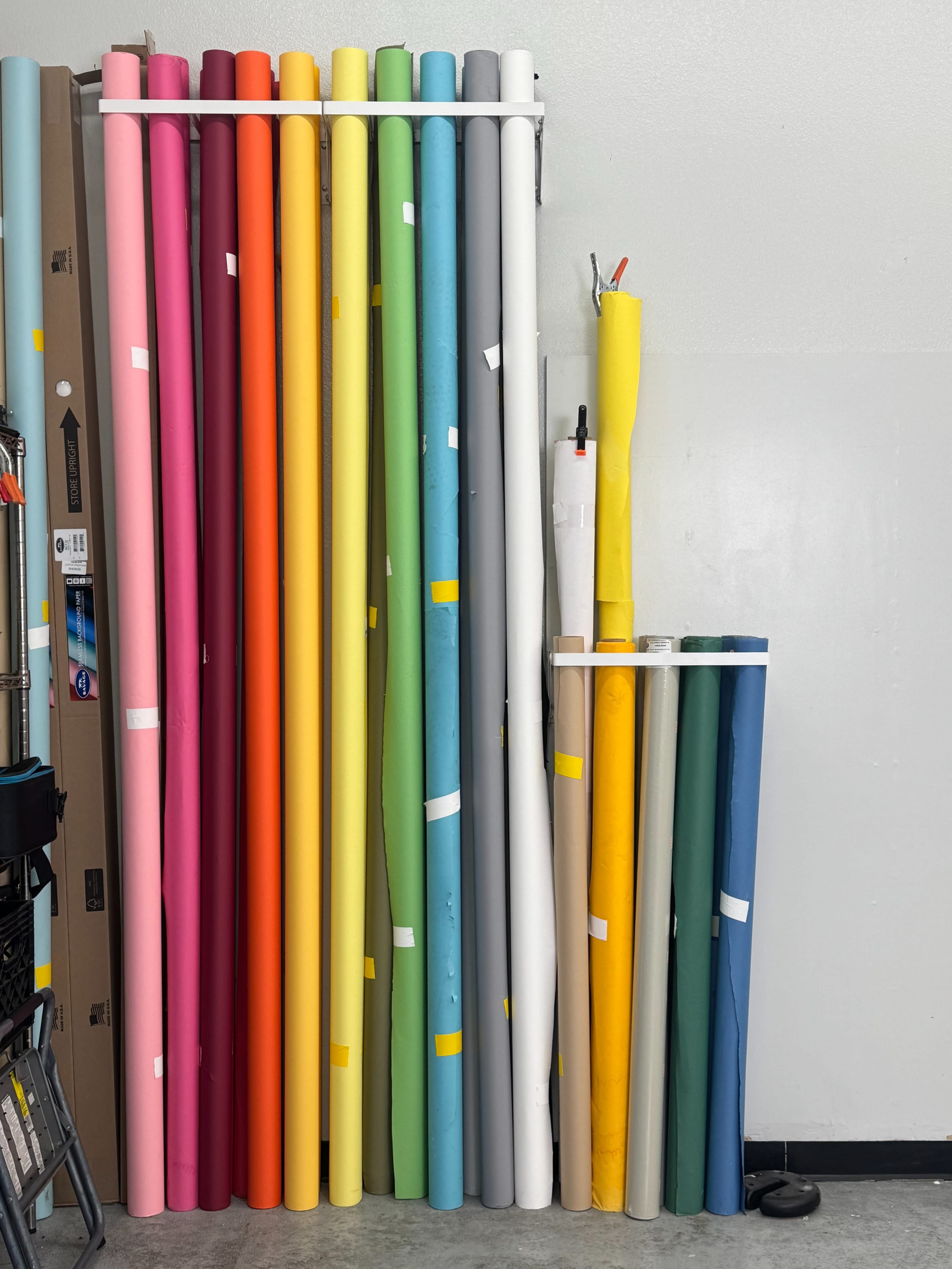 The image shows a collection of rolled sheets of colored paper organized vertically against a wall in a storage area.