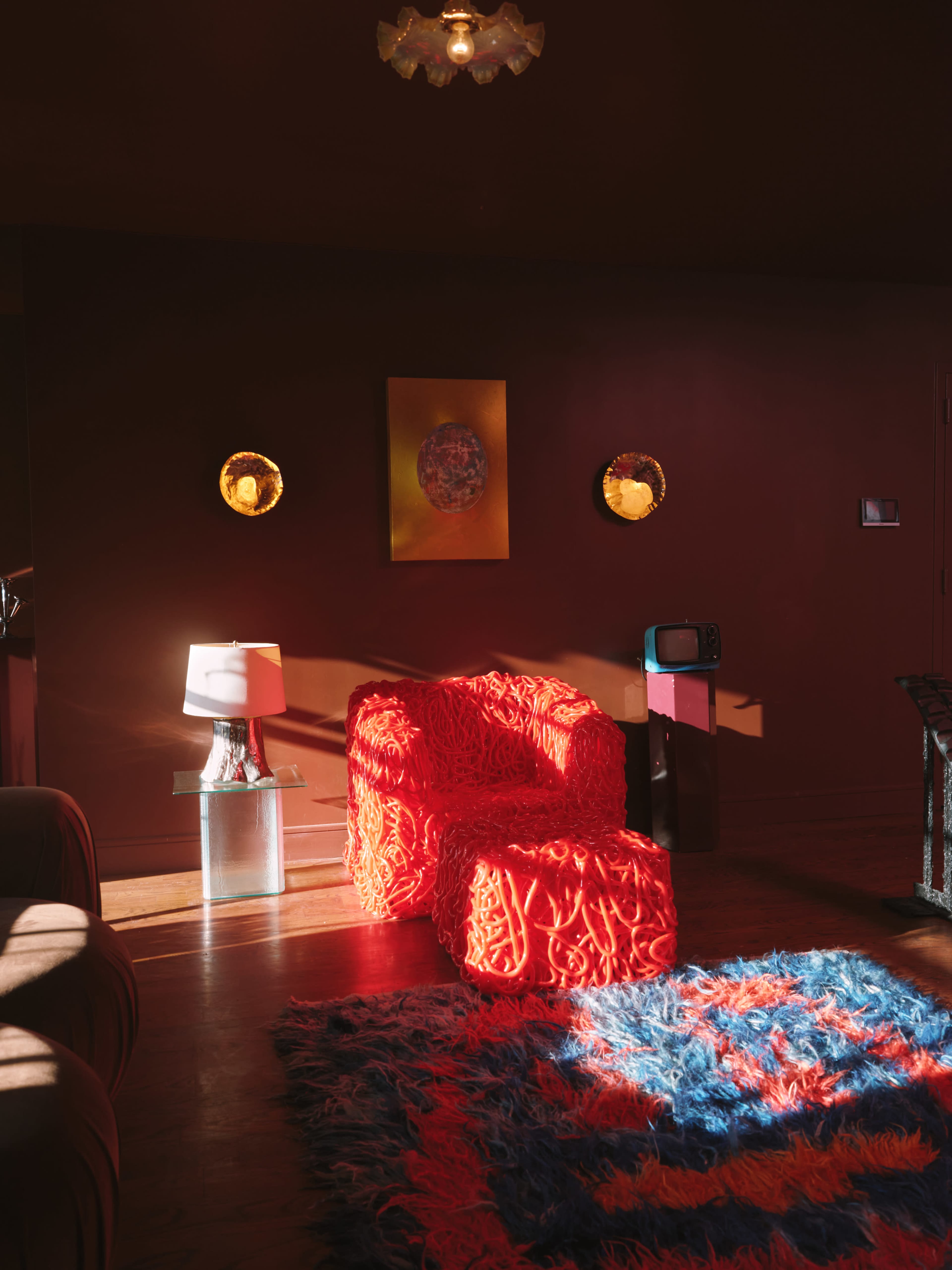 The image shows a cozy living room featuring a bright red textured armchair, a unique lamp, and a retro television, all illuminated by soft sunlight filtering through a window.