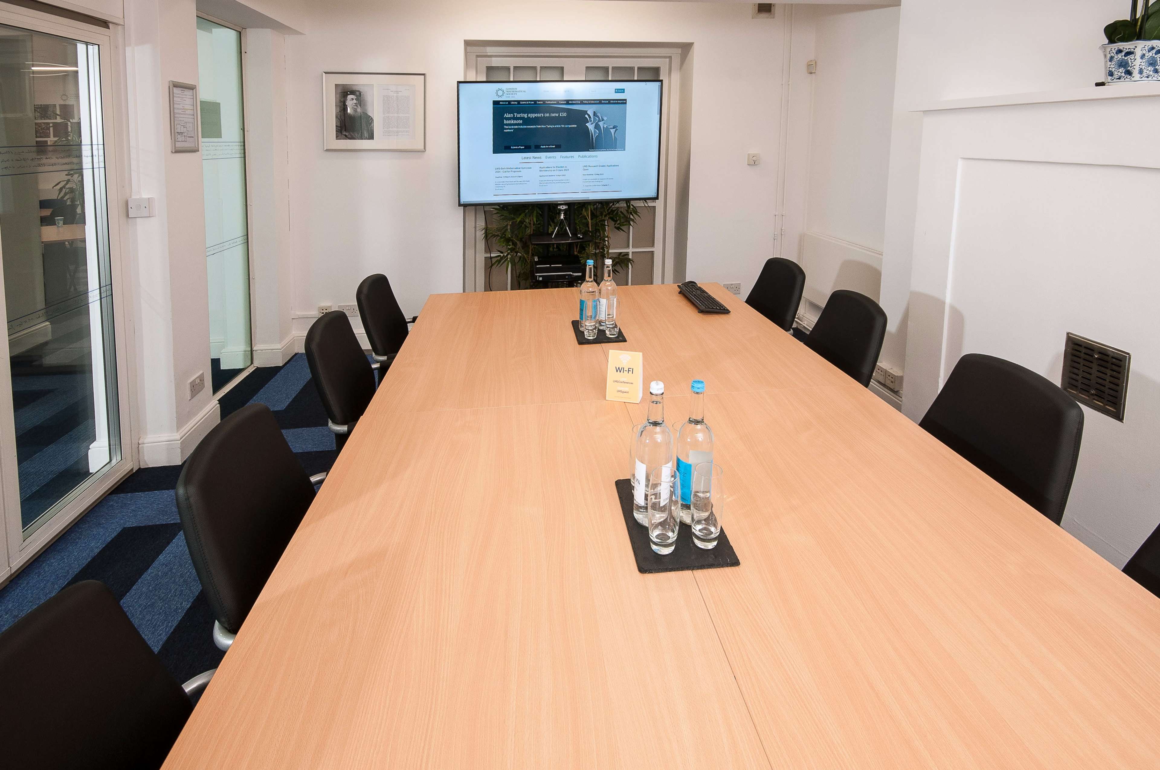 A modern conference room features a large wooden table surrounded by black chairs, with a screen displaying a website in the background.