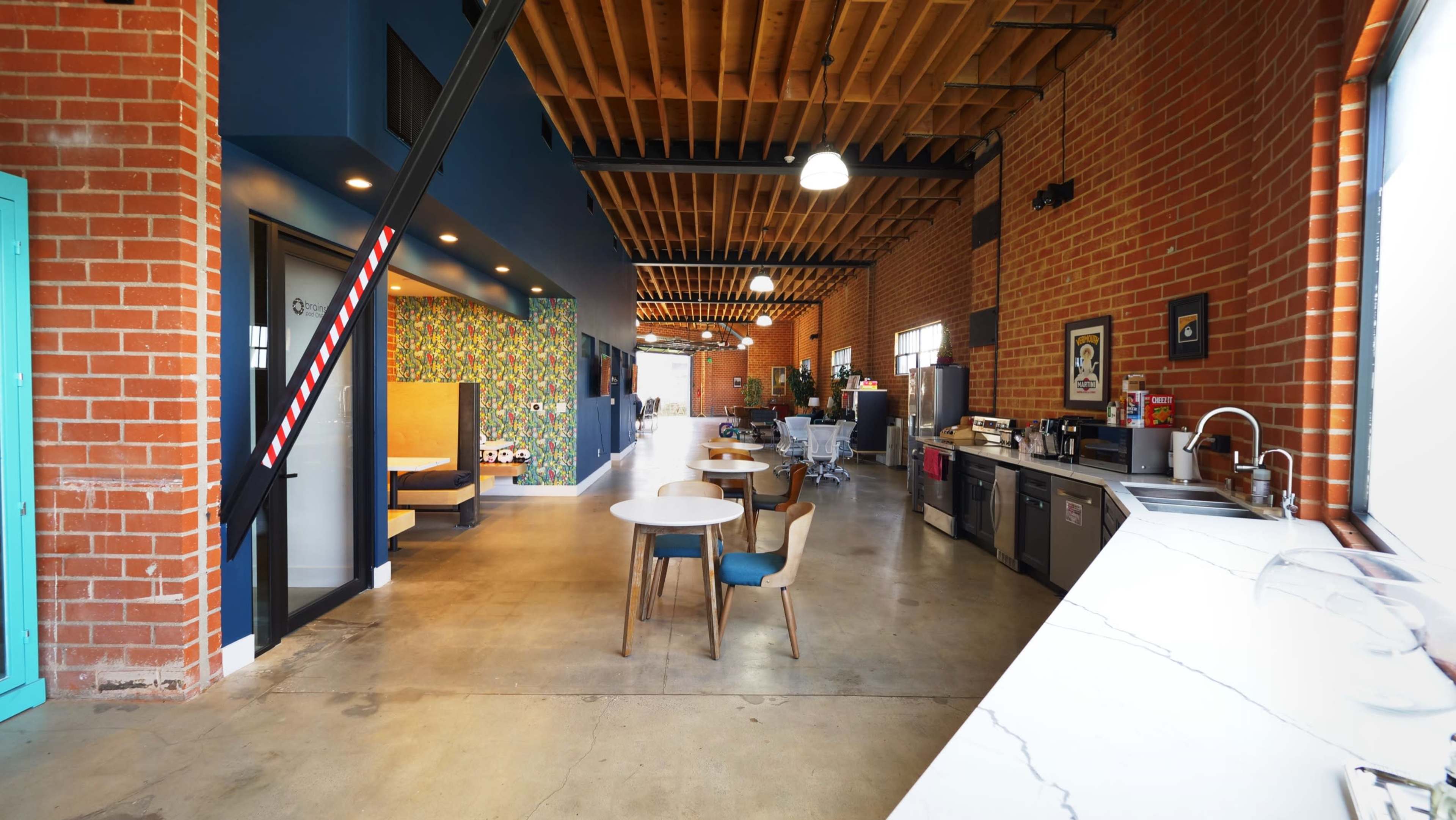 The image shows a spacious, industrial-style interior with exposed brick walls, wooden beams, a dining area featuring a round table, and a kitchen along one side.