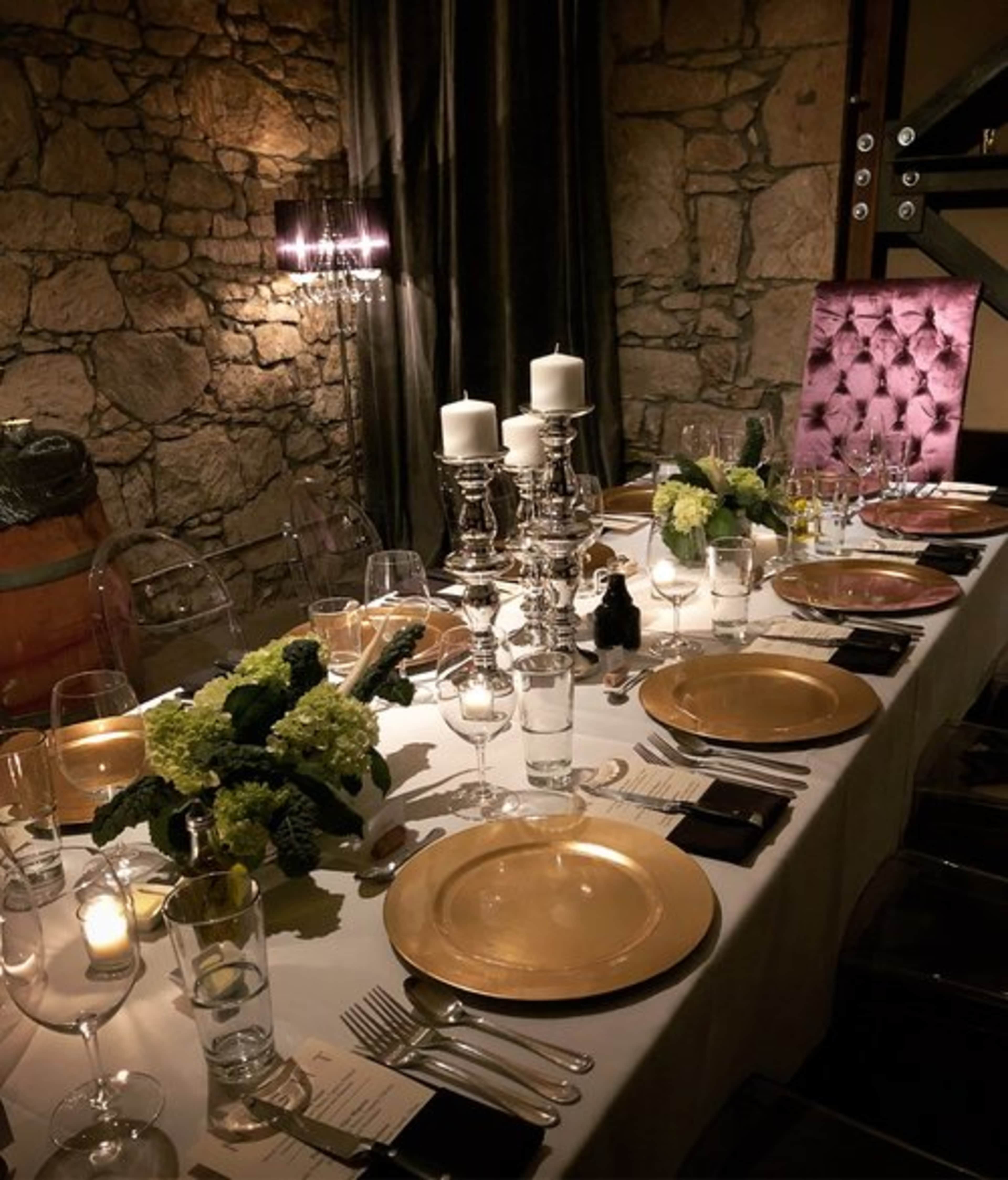 A elegantly set dining table features gold charger plates, glassware, candles, and floral arrangements against a stone wall backdrop.