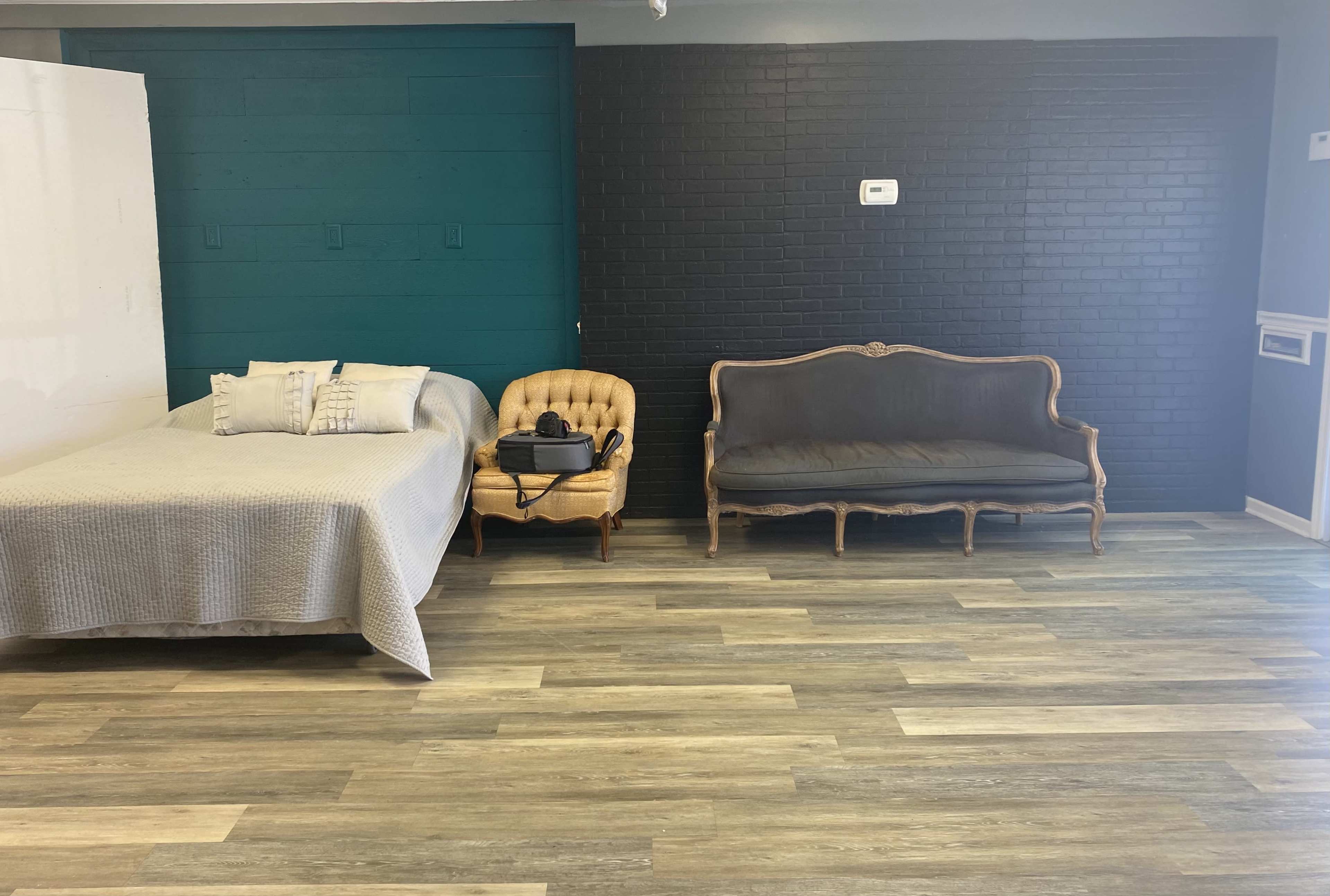 A room features a bed with a light cover, a vintage armchair, and a dark upholstered sofa against a black brick wall.