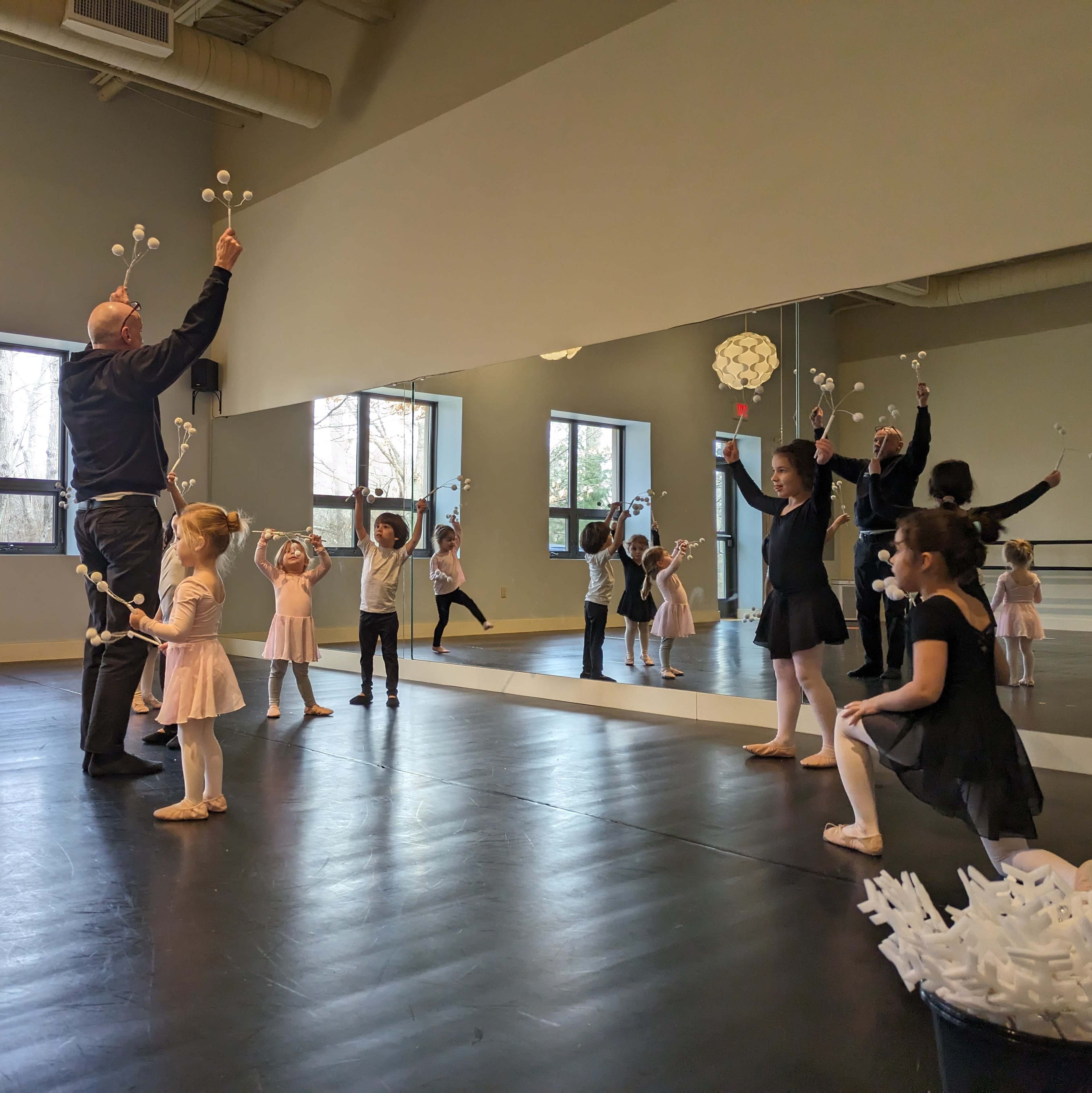 A group of young dancers is practicing in a studio, holding props while an adult instructor leads them.