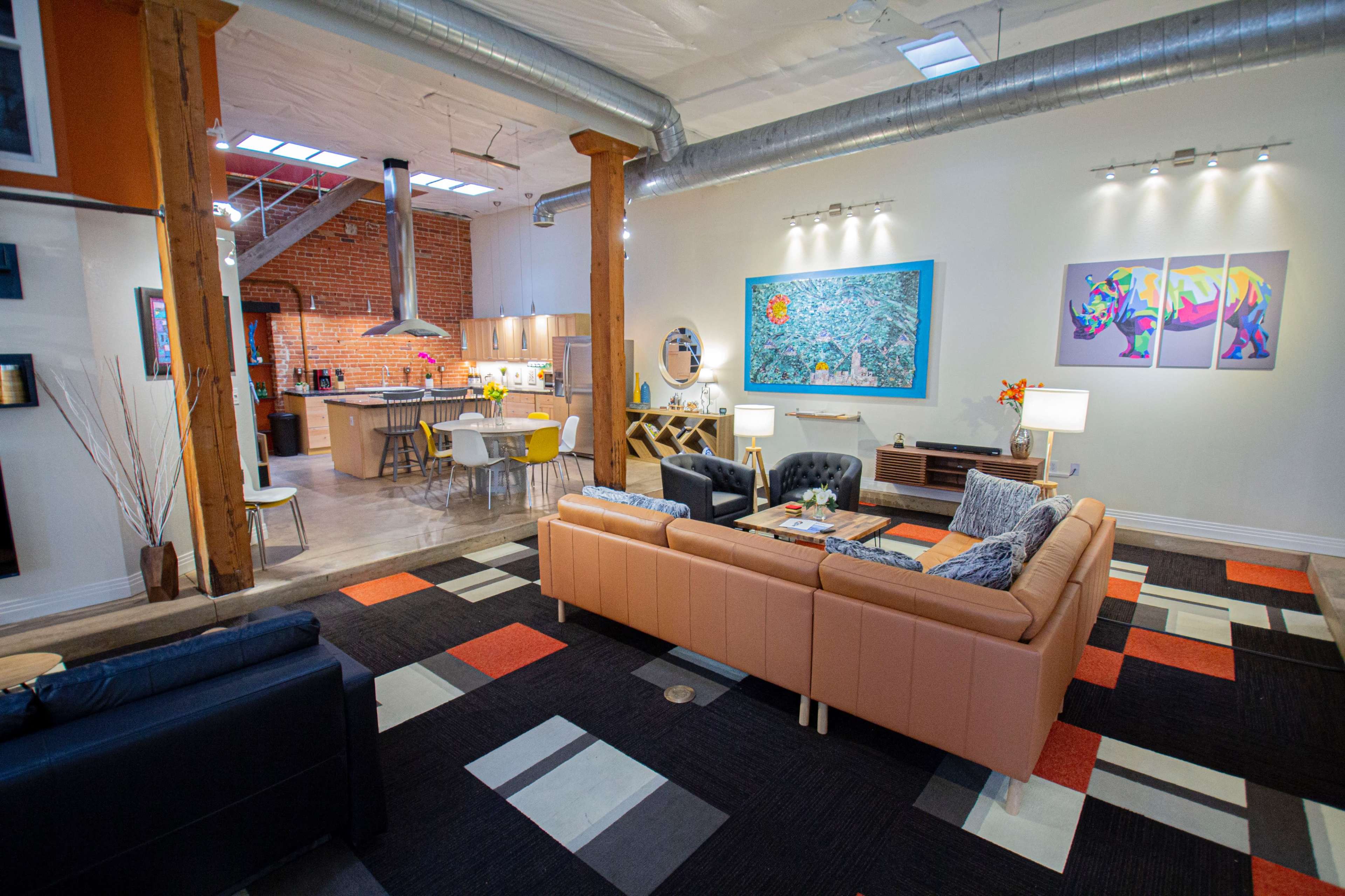 The image shows a spacious, modern loft with a living area featuring a sofa, coffee table, and colorful area rugs, adjacent to an open kitchen with brick walls and a dining space.