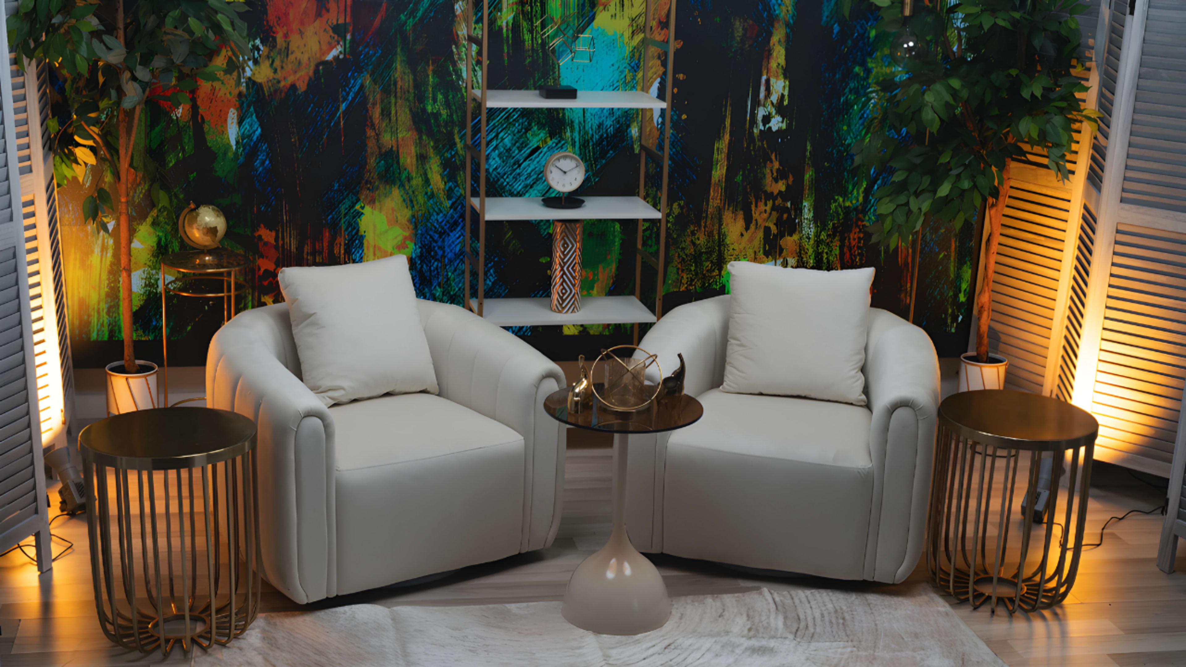 The image shows a modern living room setup with two white armchairs, a round table between them, and a colorful abstract backdrop decorated with plants and ambient lighting.
