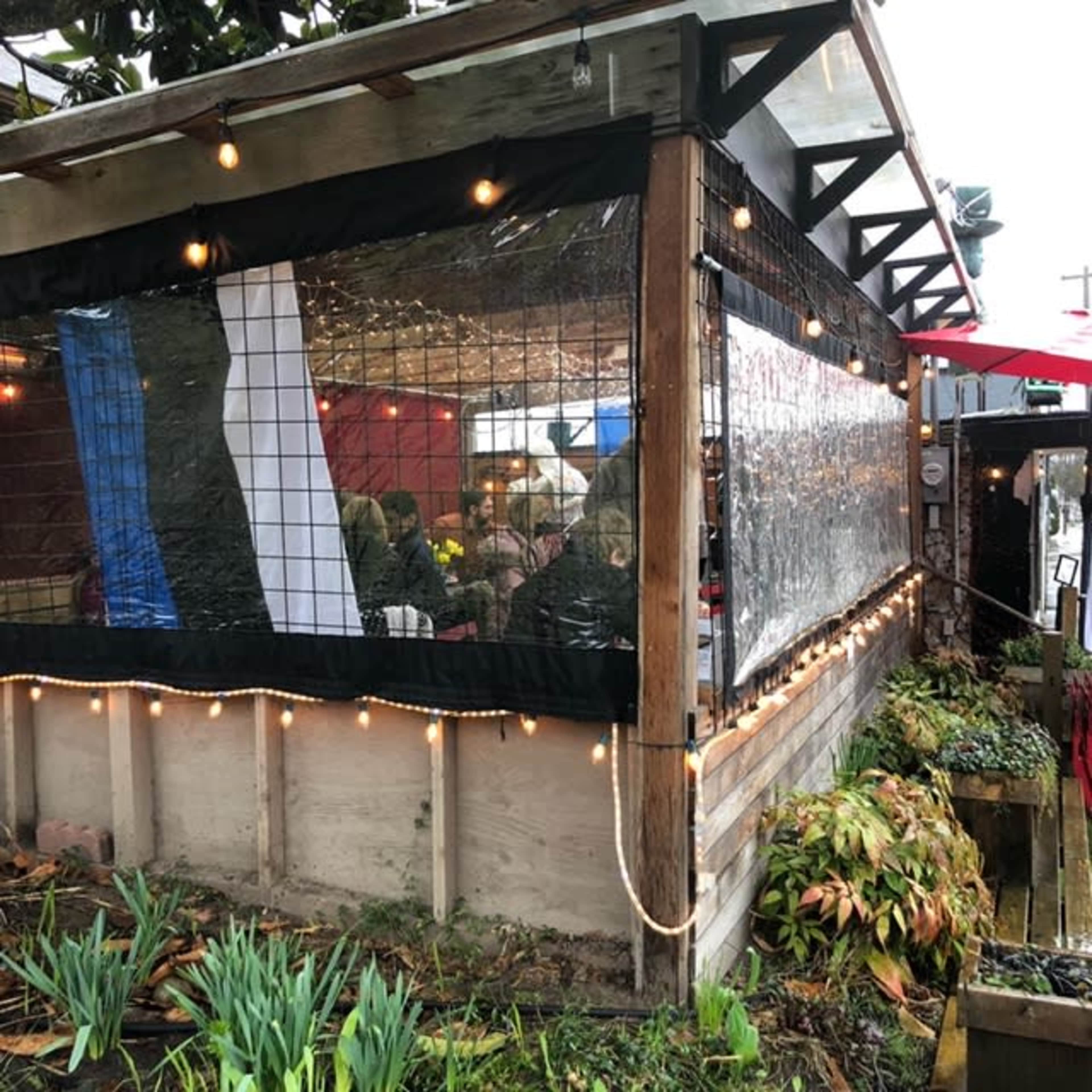 A covered outdoor dining area with string lights and tables is visible, surrounded by greenery and featuring a transparent plastic barrier.