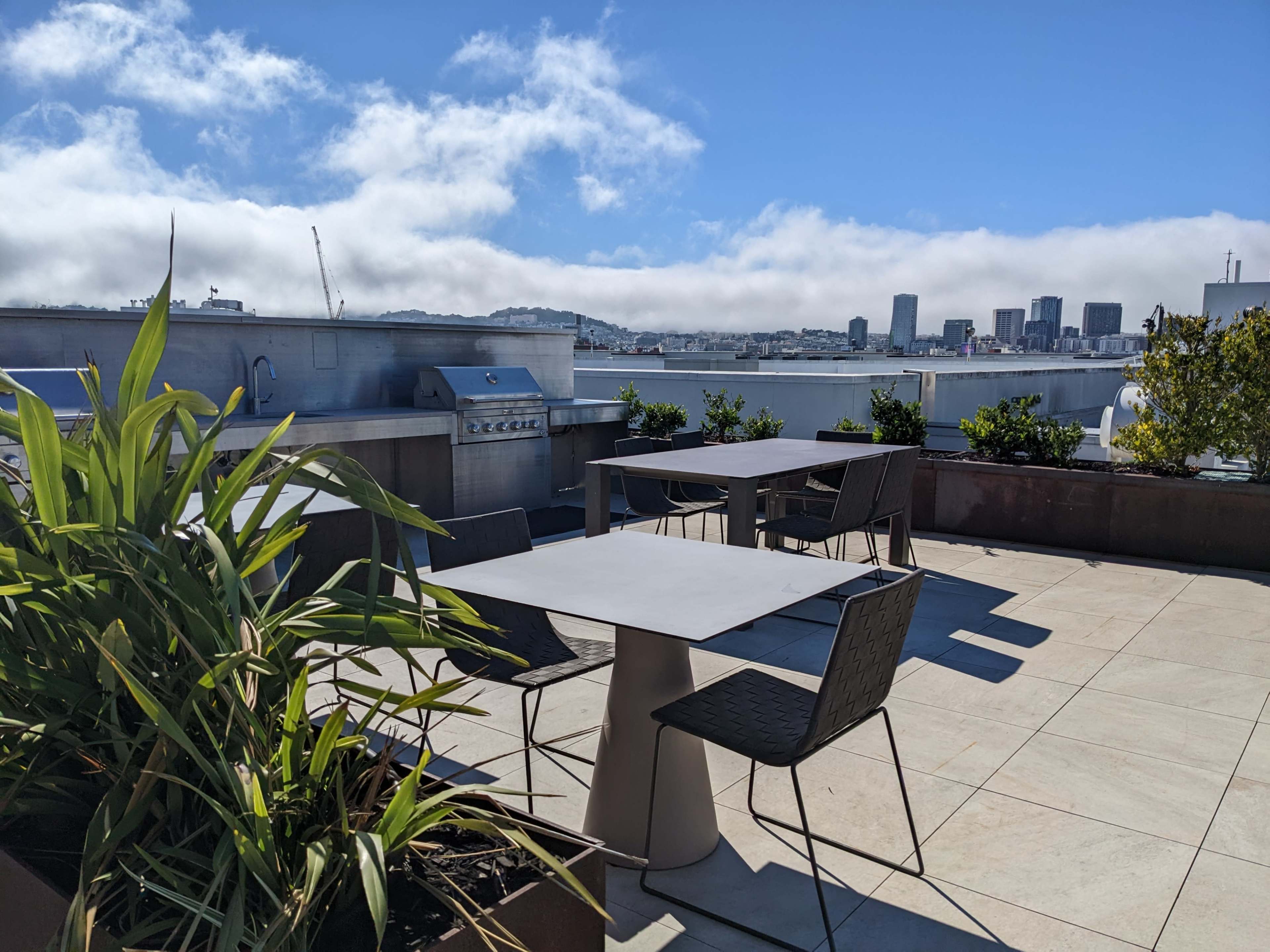 A rooftop patio with tables and chairs, a barbecue grill, and a view of a city skyline under a partly cloudy sky.