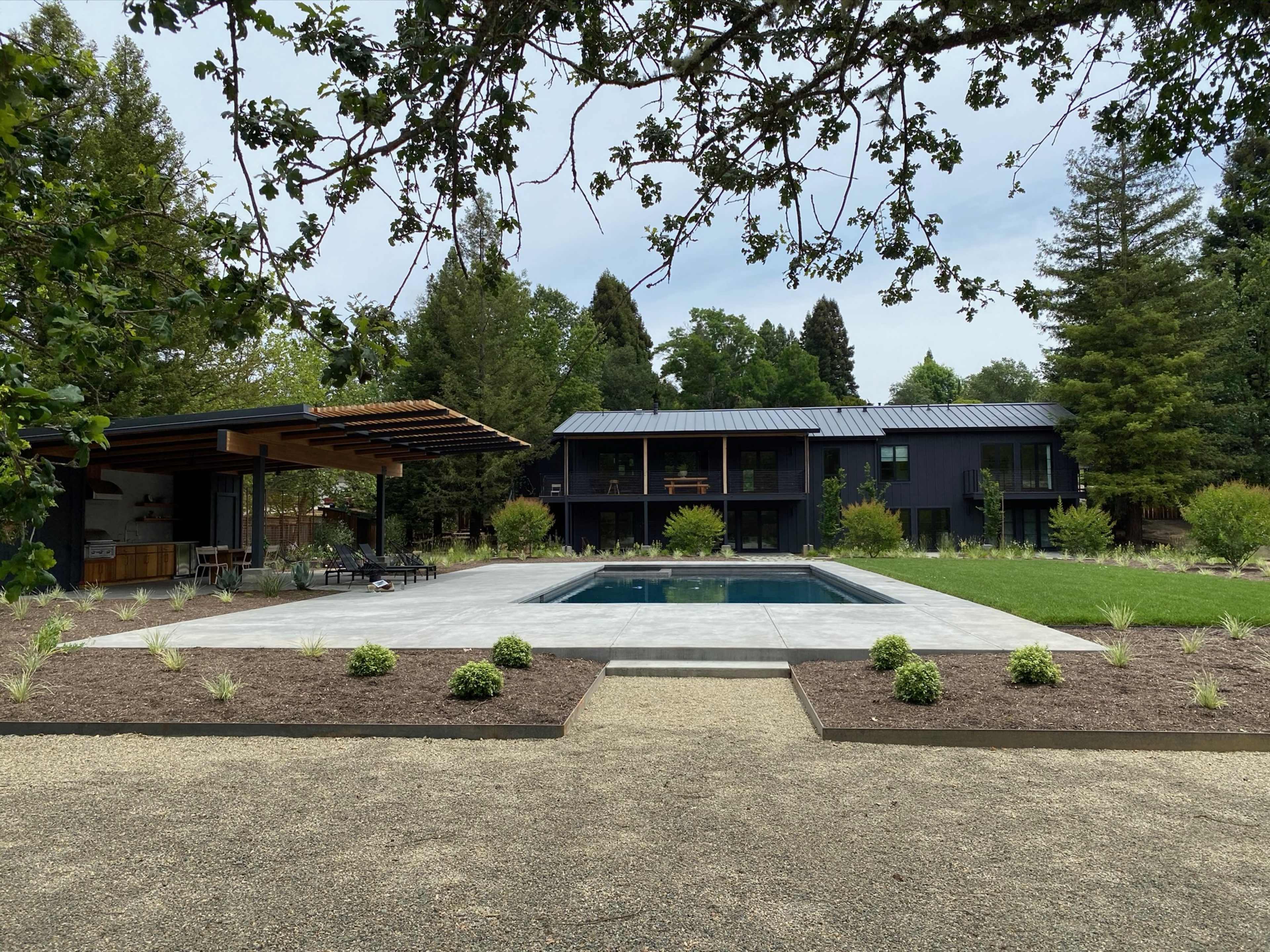 A modern two-story black house is situated beside a rectangular swimming pool, surrounded by landscaped greenery and a patio with outdoor seating.