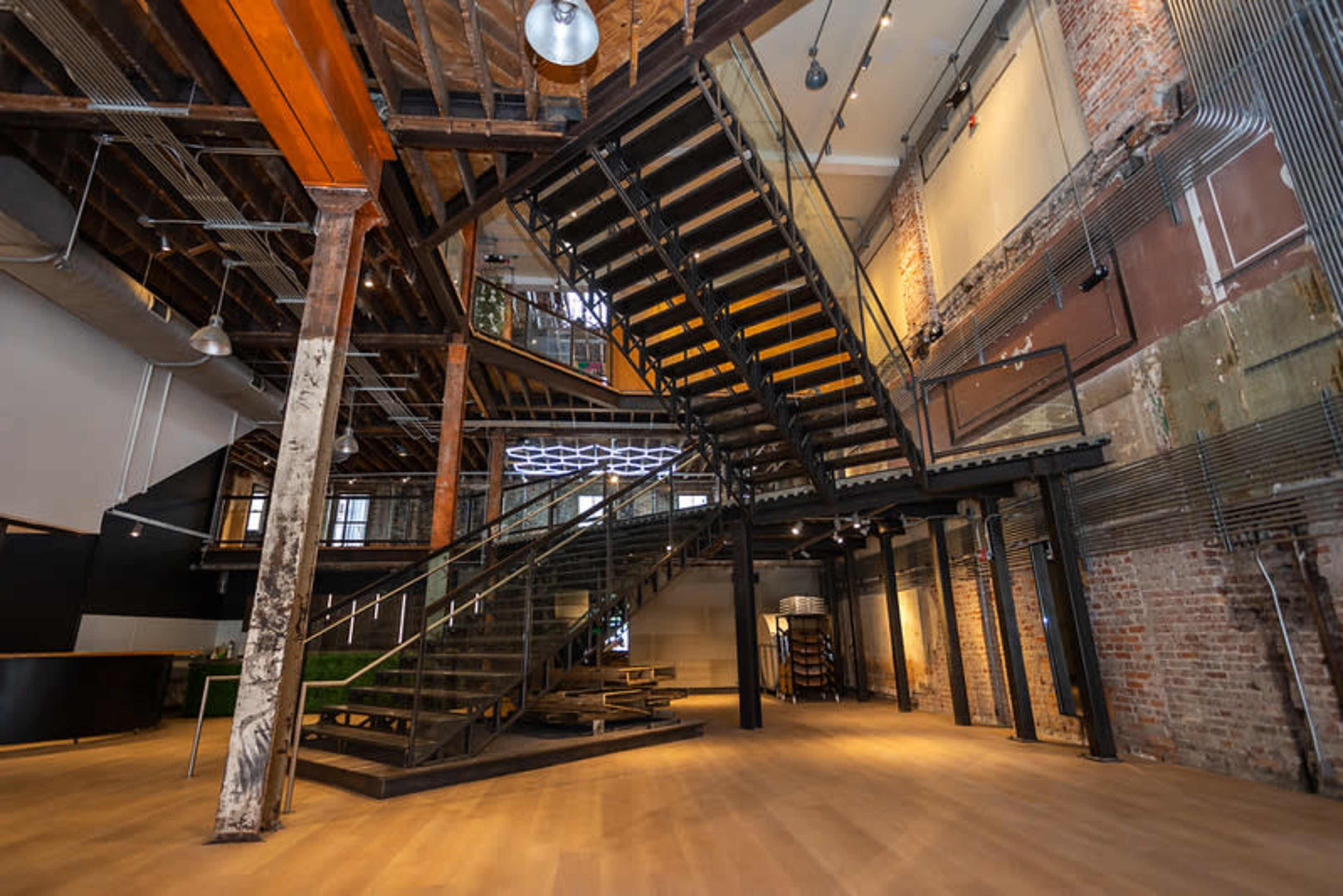 The image shows a modern interior space featuring a prominent staircase made of metal and wood, surrounded by exposed brick walls and industrial design elements.