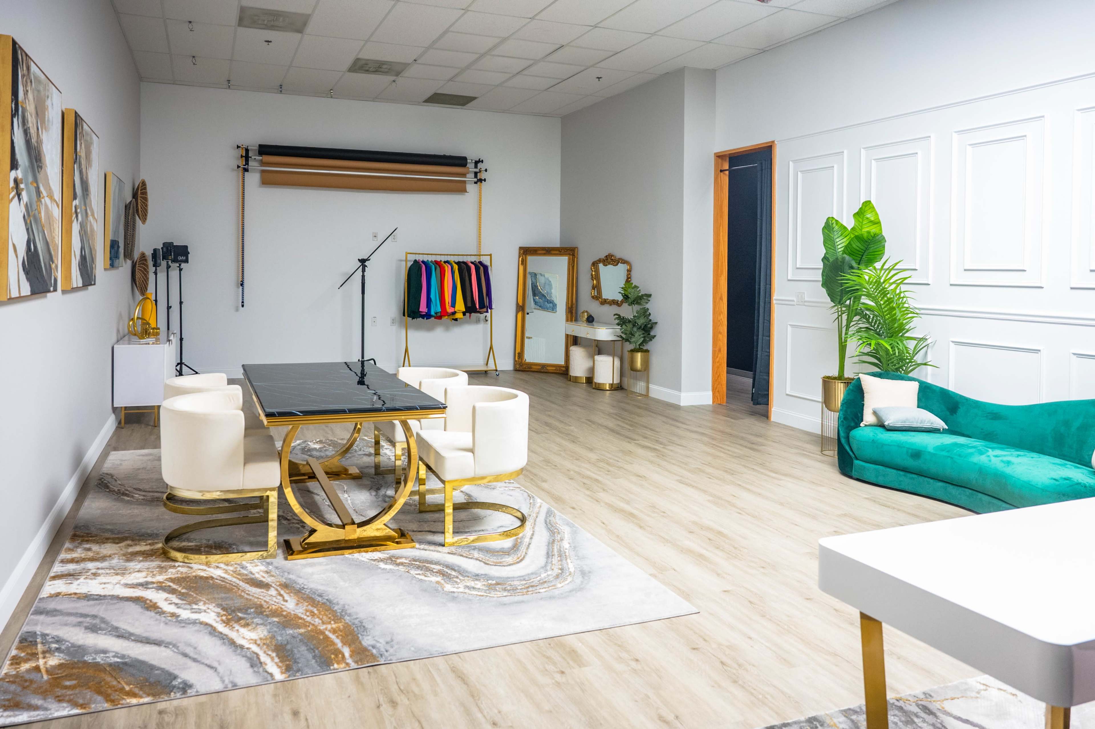 A spacious interior room features a dining table with four chairs, a sofa, and a wall with artwork, along with clothing on hangers and decorative plants.