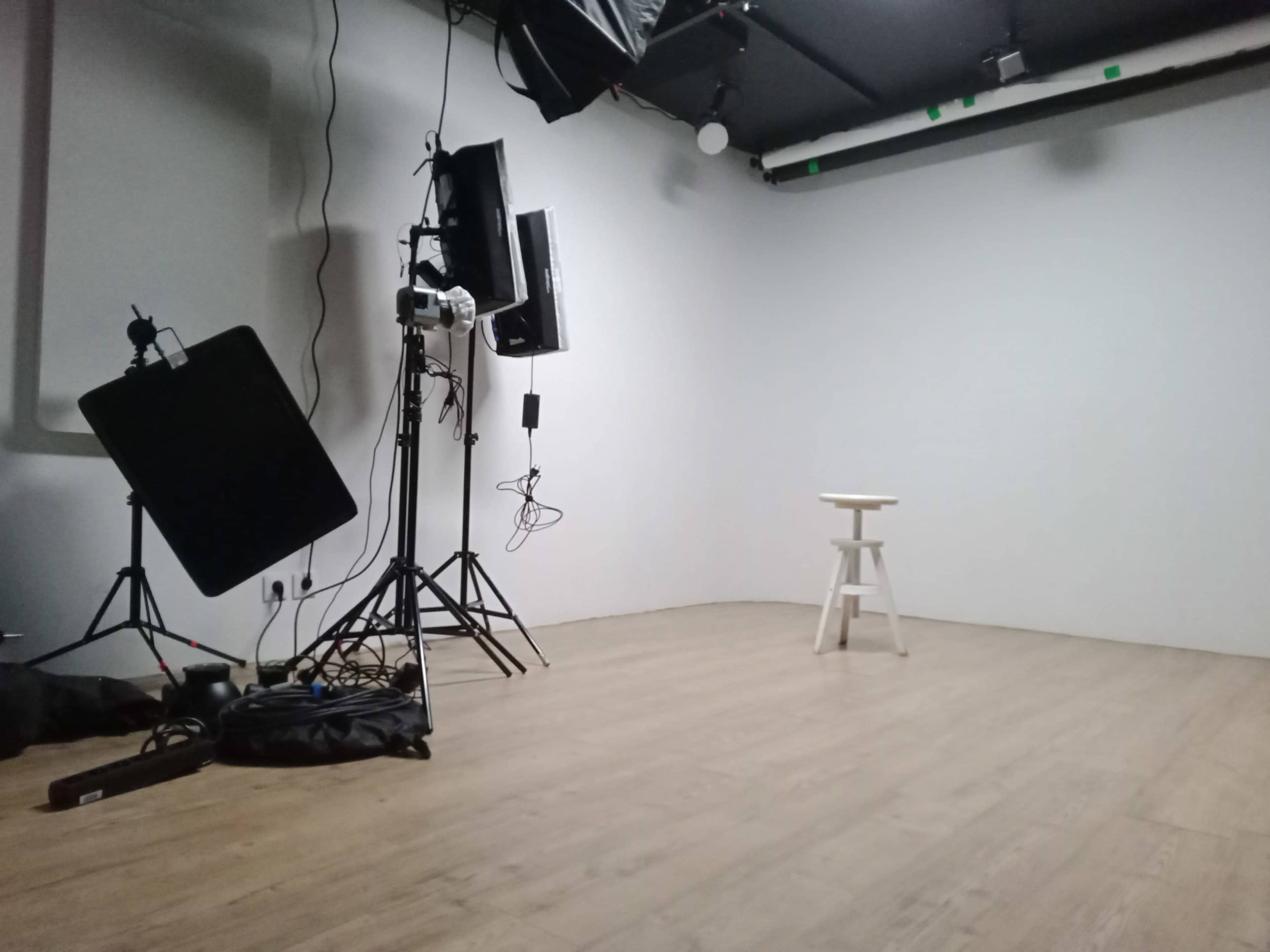 The image shows a blank studio space with a white wall, a wooden floor, and a single white stool in the center, surrounded by lighting equipment on stands.