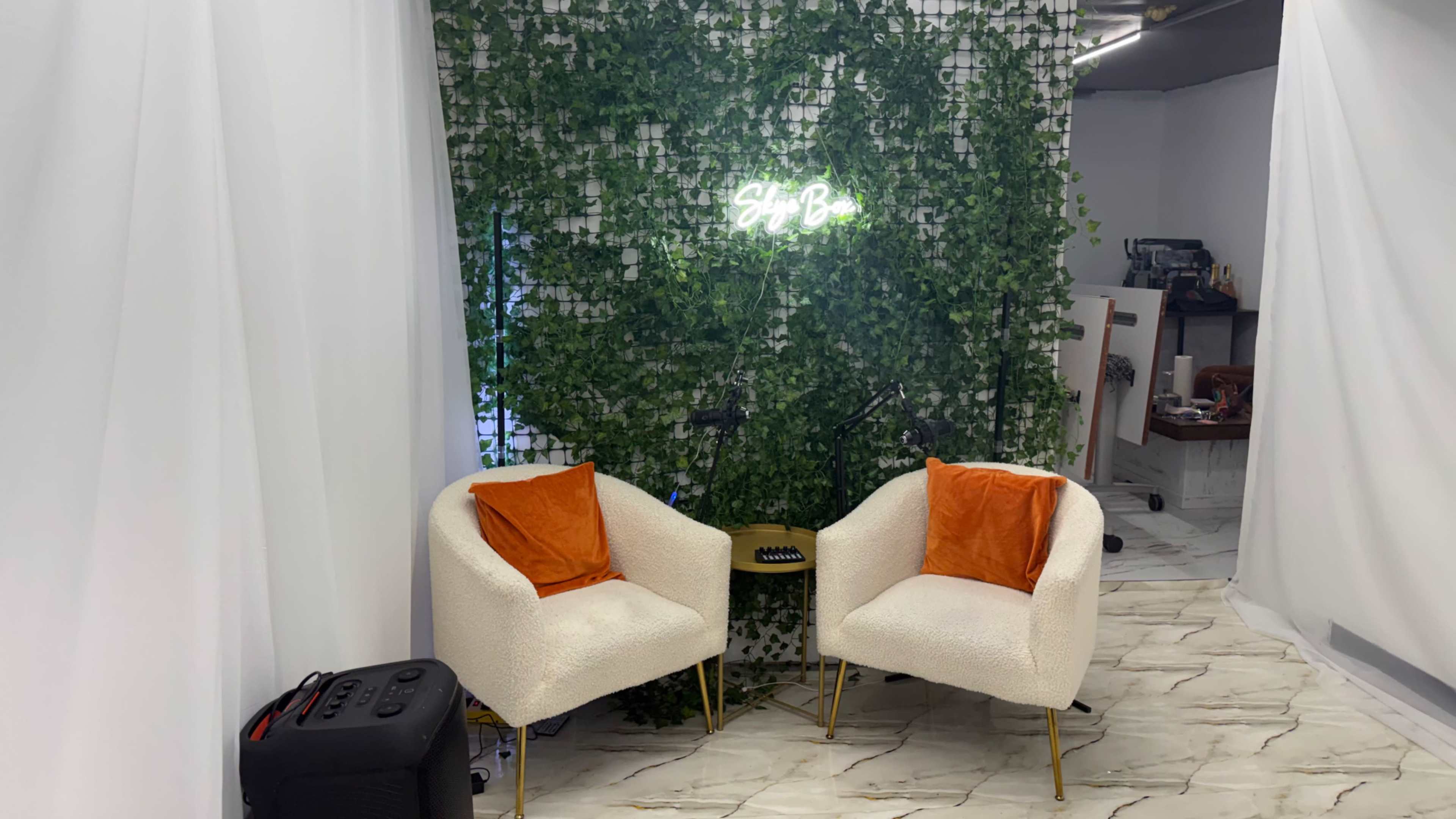 The image features a small seating area with two white chairs adorned with orange cushions, set against a green wall covered in foliage and illuminated by a neon sign.