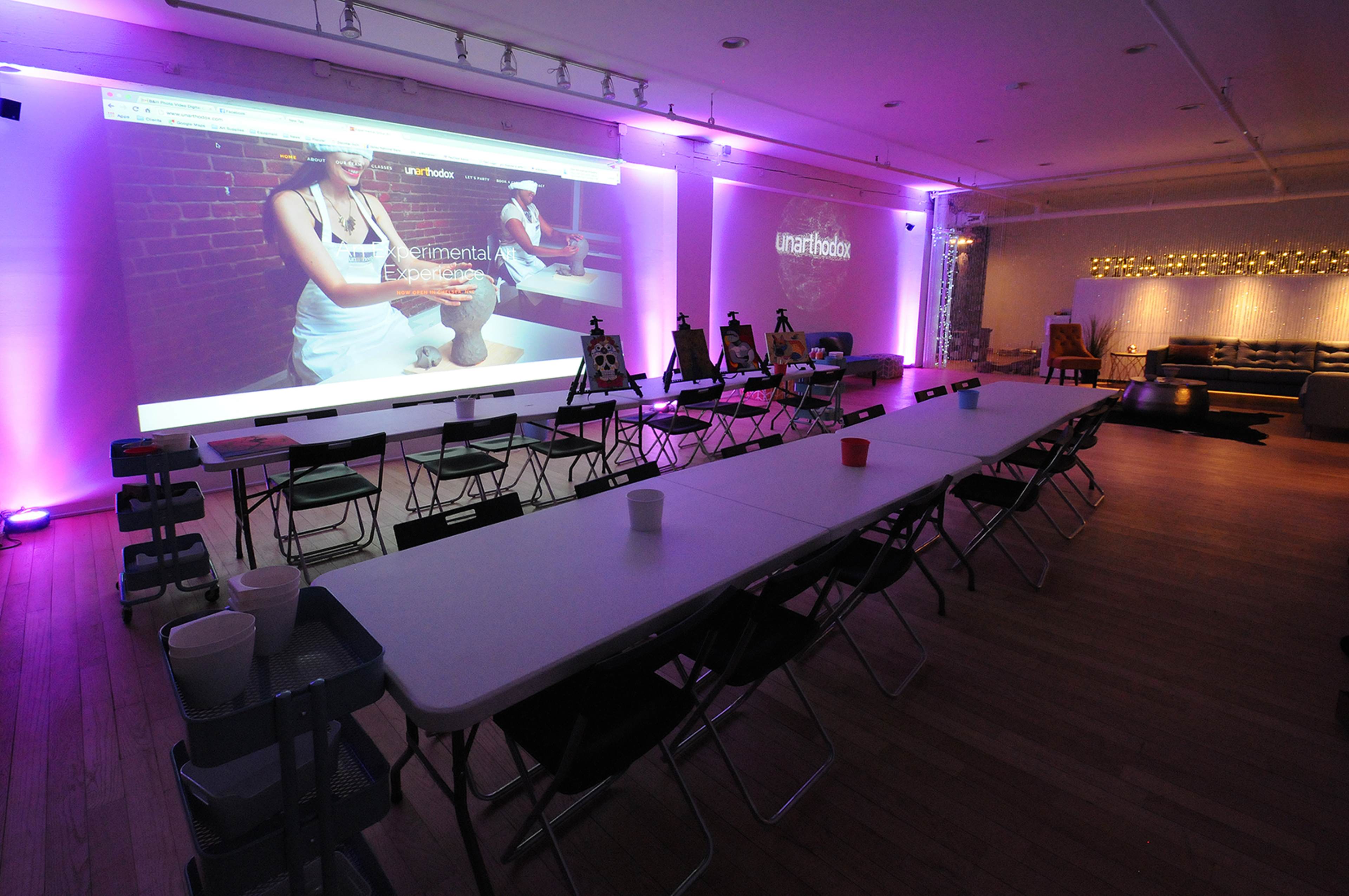 A spacious room is set up with a long table and chairs, featuring a large projector screen displaying a website, illuminated by purple lights.