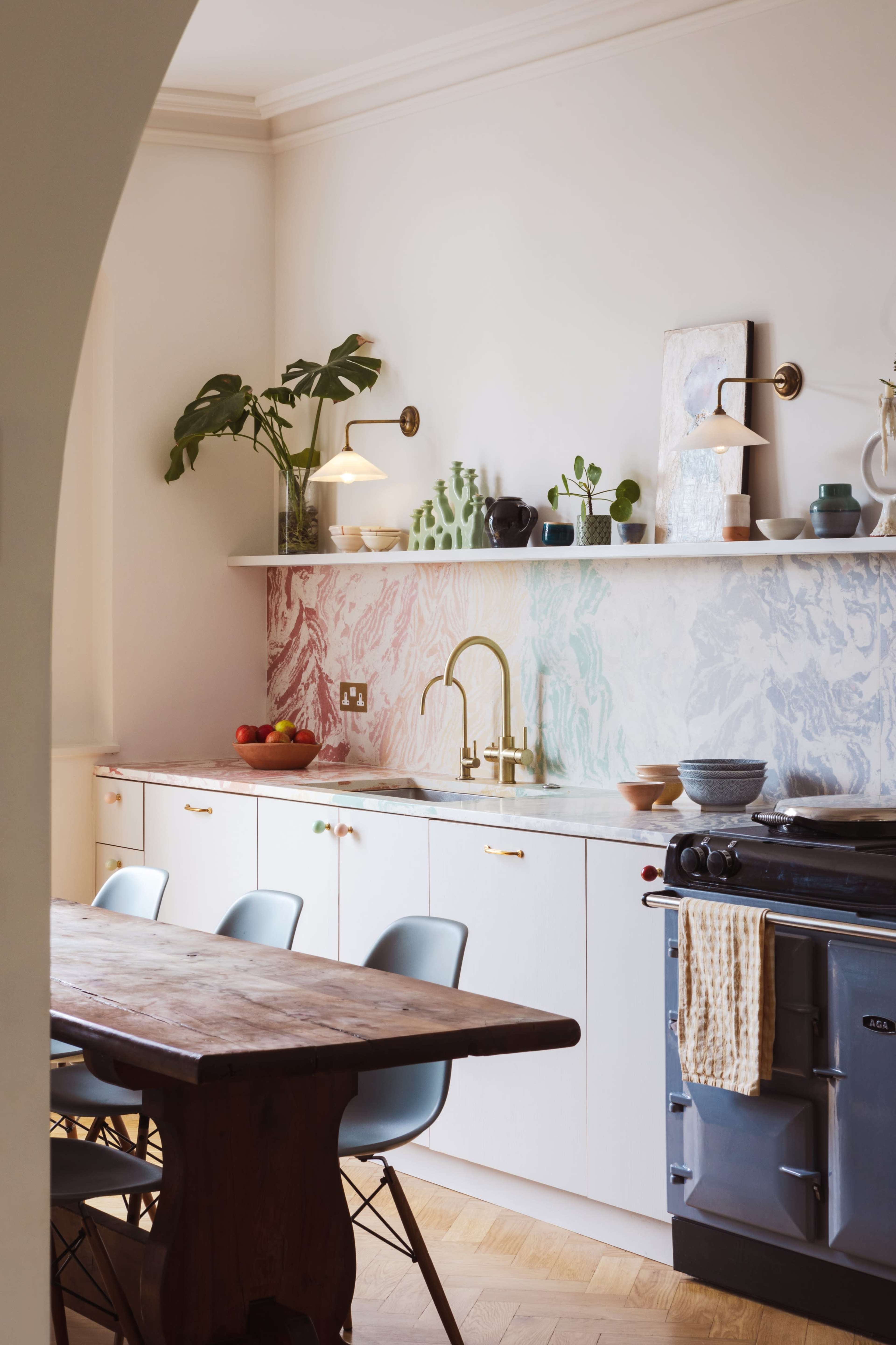 A modern kitchen features a marble-patterned backsplash, a wooden dining table, and a brass faucet above a cream-colored countertop.