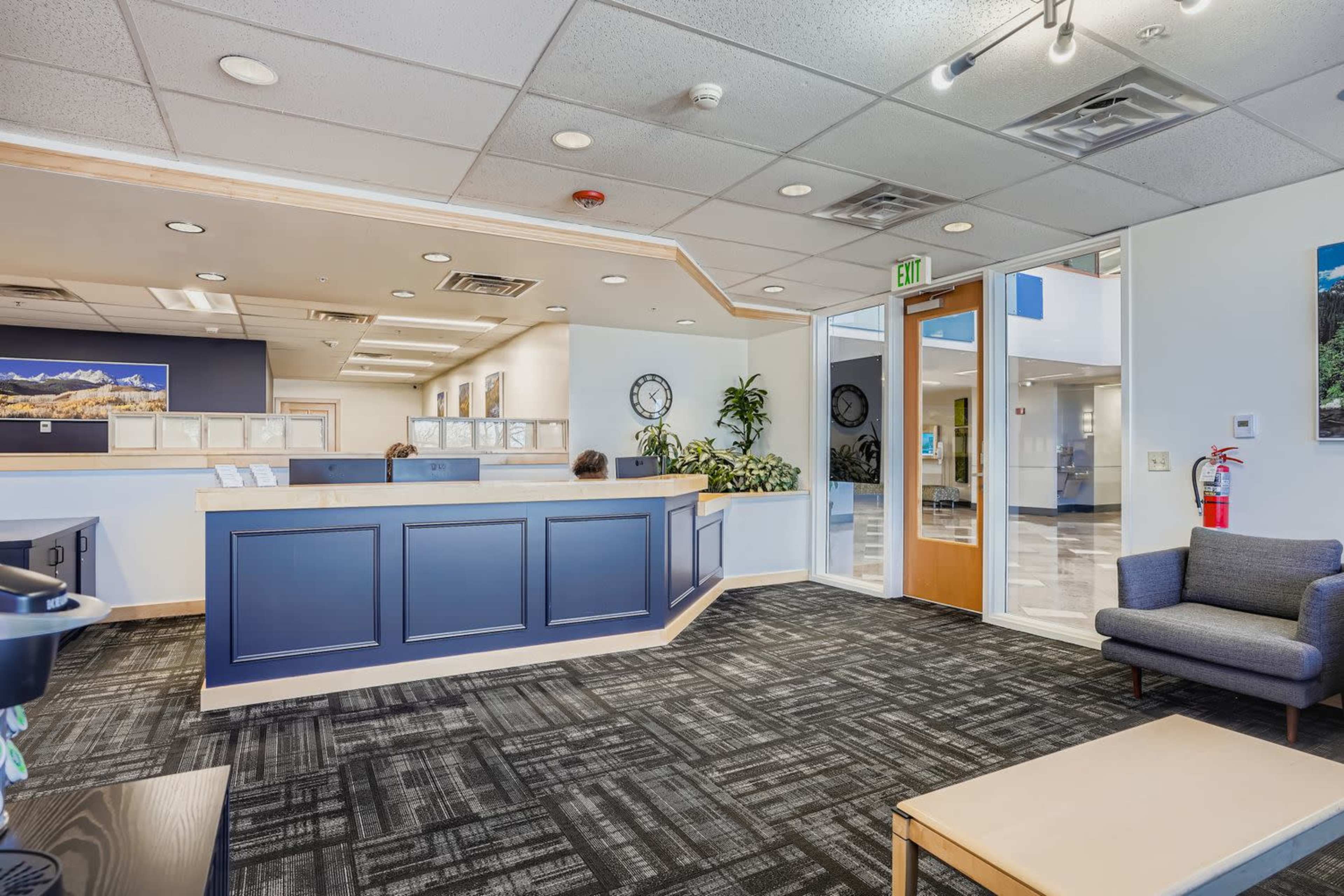The image shows a modern reception area with a blue front desk, gray seating, and large windows letting in natural light.