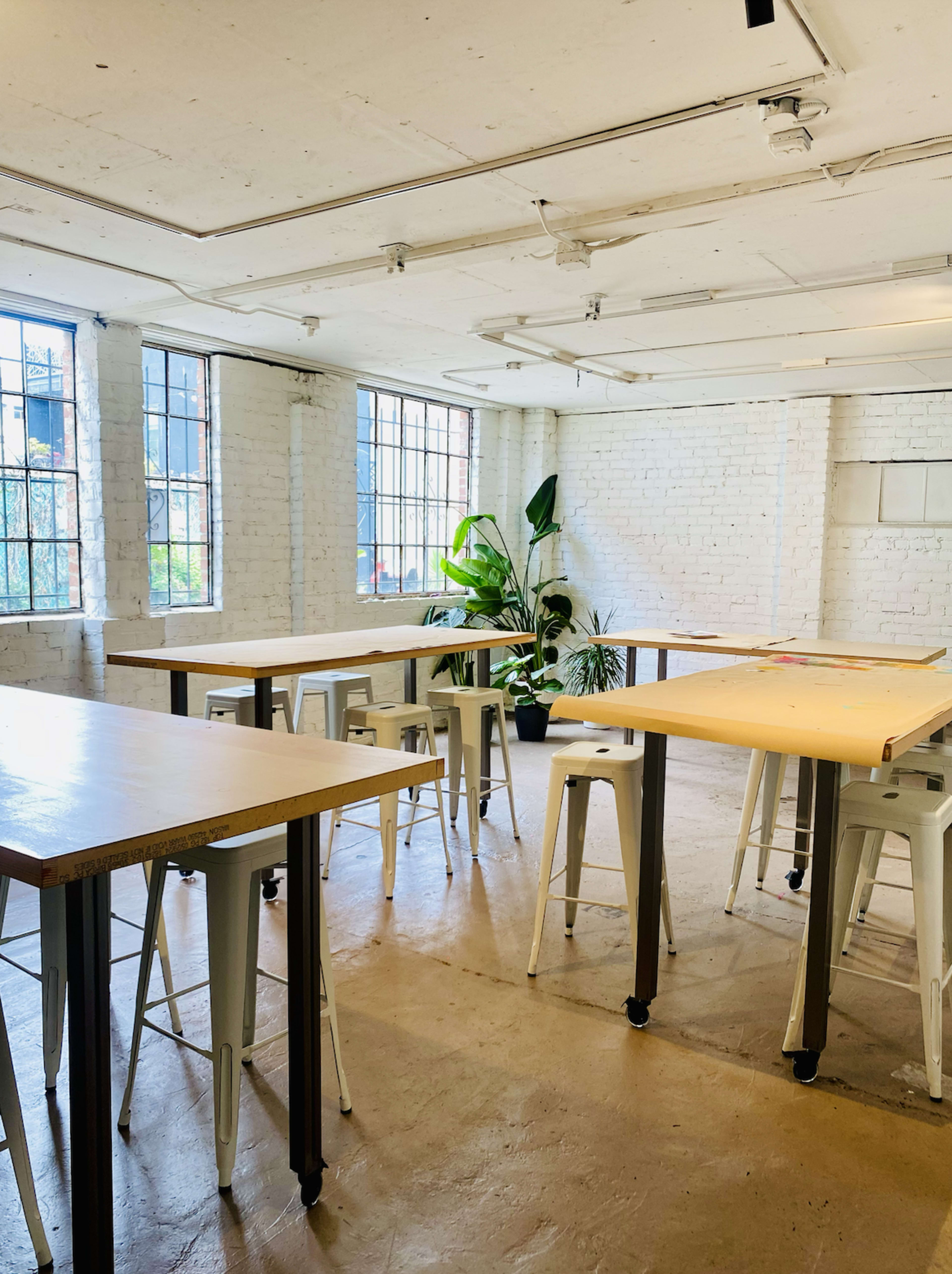 A spacious room with large windows, featuring several tall tables and metal stools, along with a potted plant in the corner.