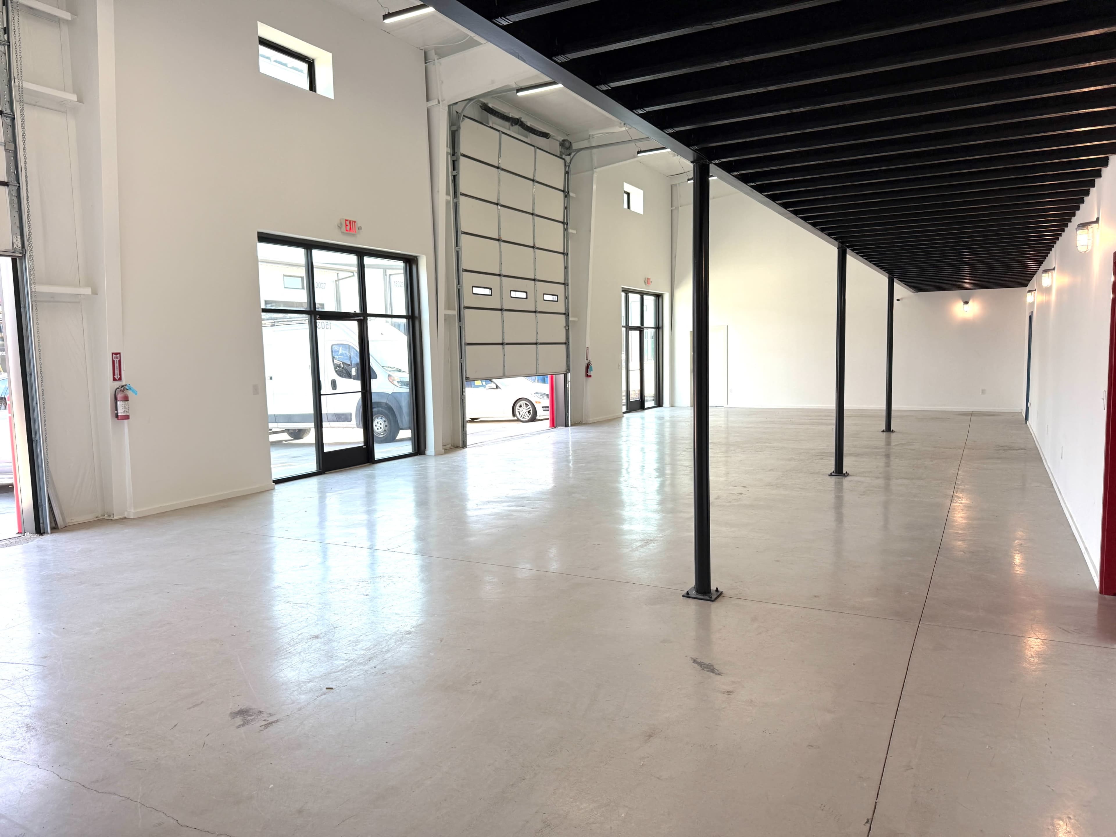 The image shows an empty industrial space with high ceilings, large windows, and a garage door at one end.