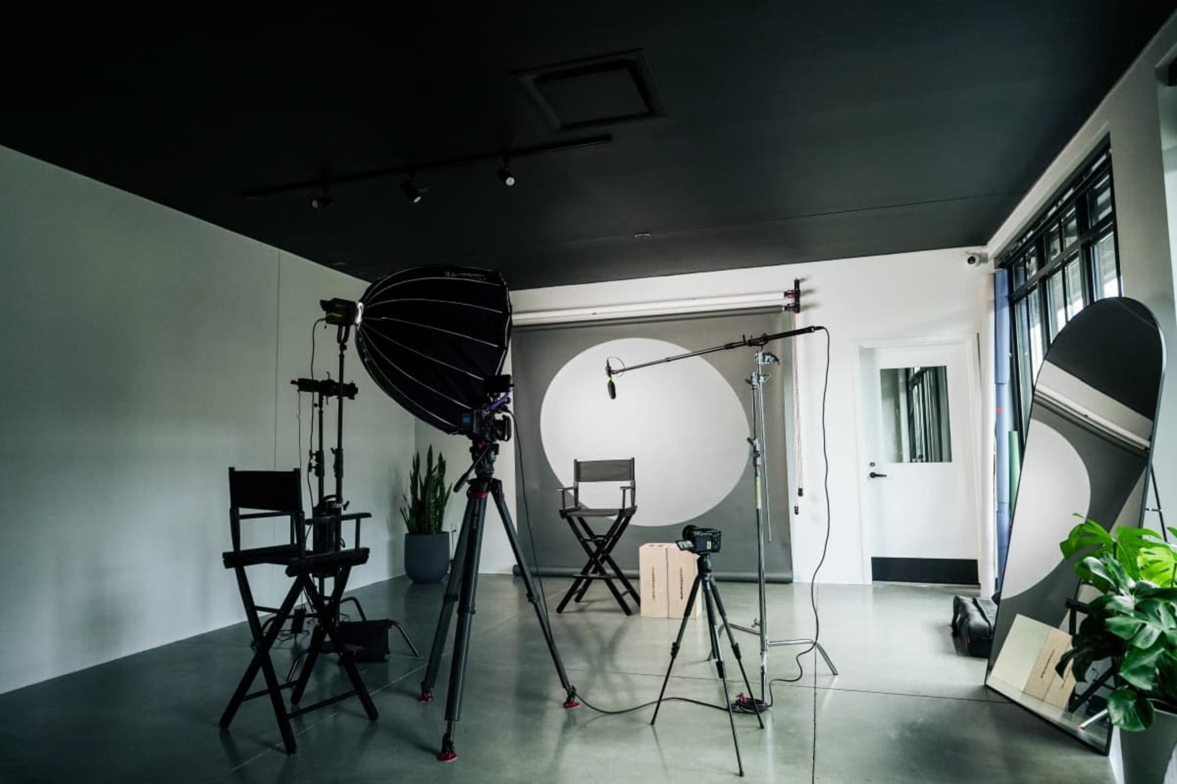 The image shows a photography studio equipped with lights, cameras, and empty director's chairs, featuring a circular backdrop and plants.
