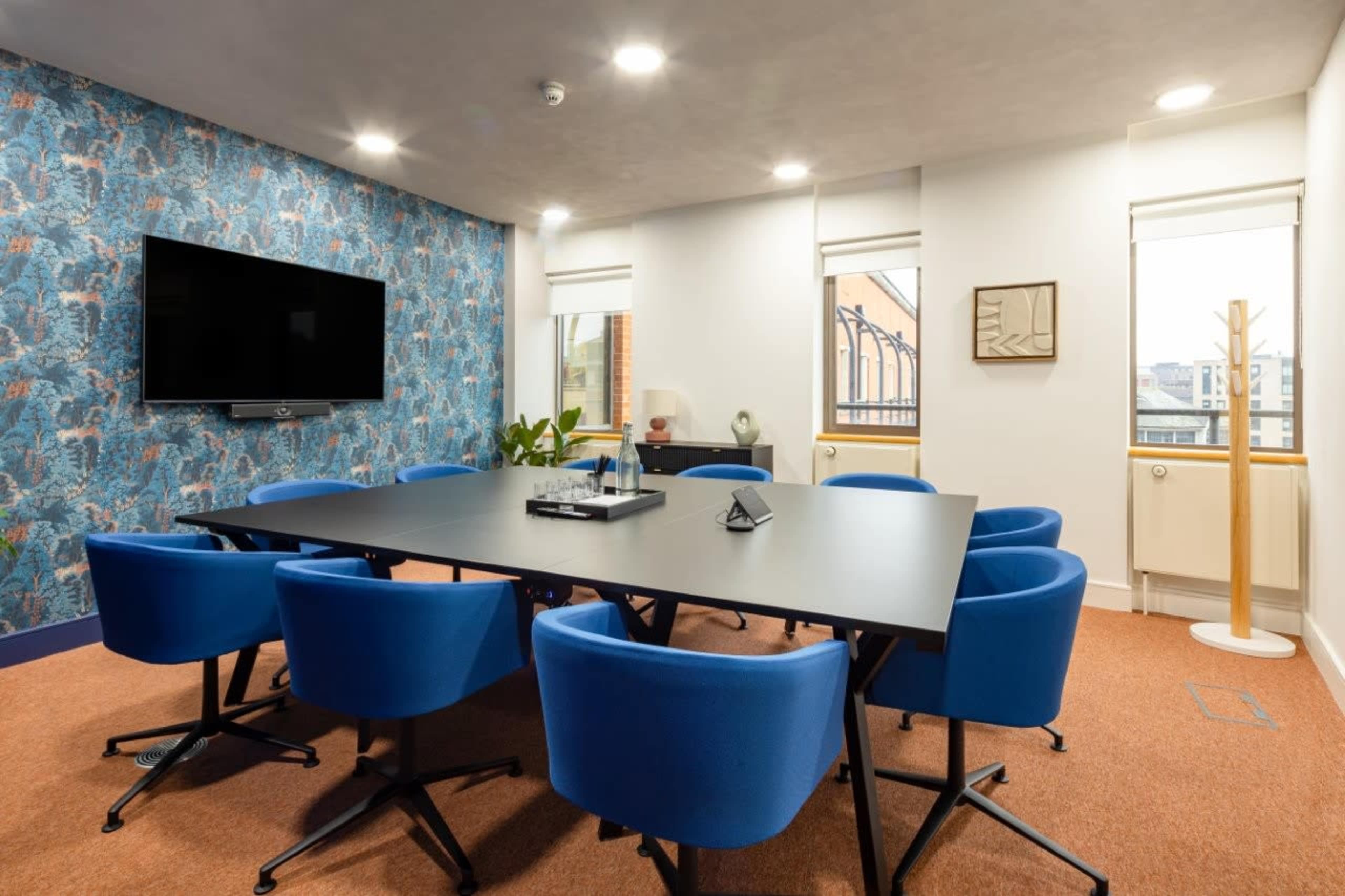 A modern conference room features a large black table surrounded by blue chairs, with a patterned wall and a television screen mounted on one side.