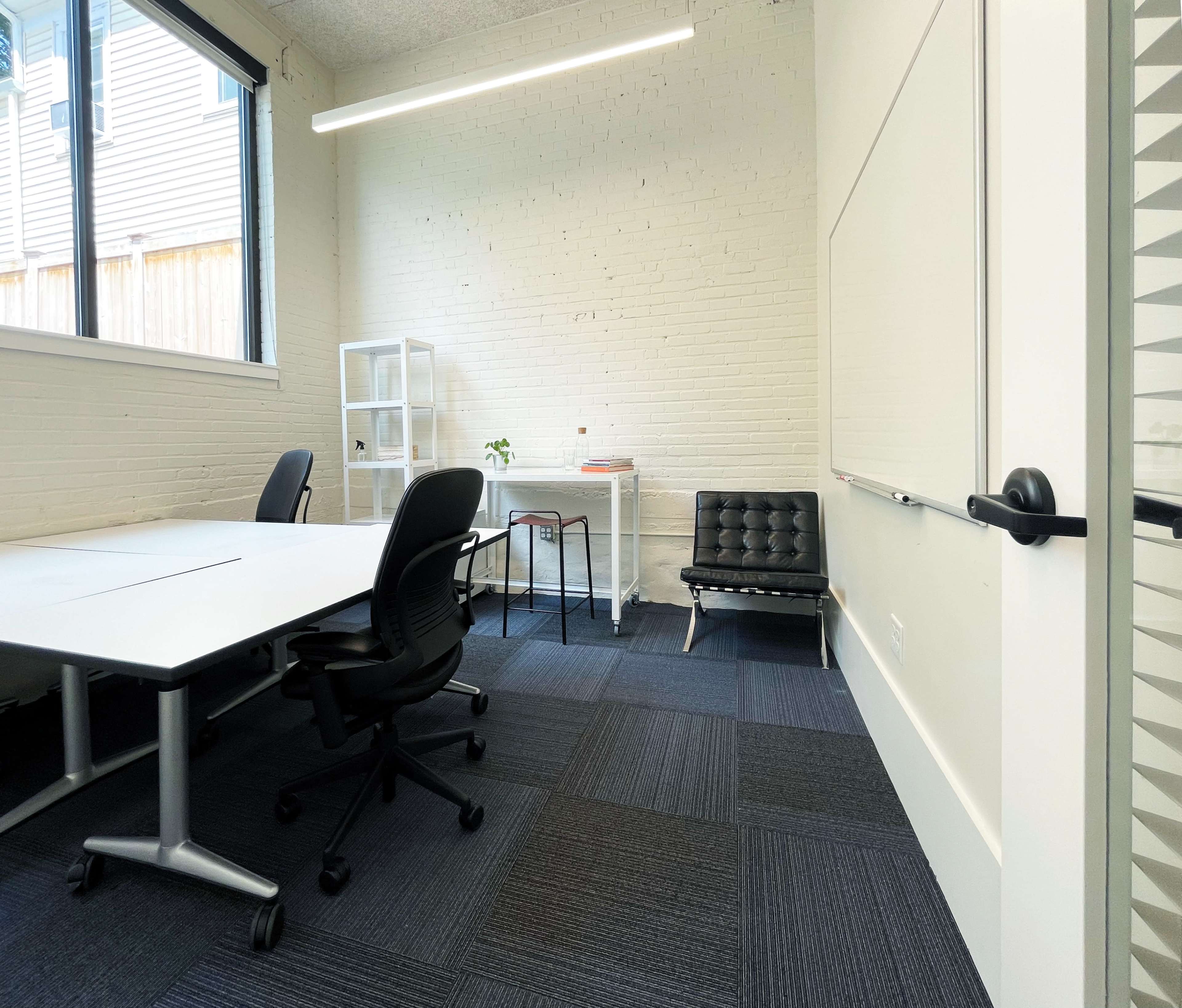 A bright, minimalistic office space featuring a large table, black chairs, a whiteboard, and natural light coming through the windows.