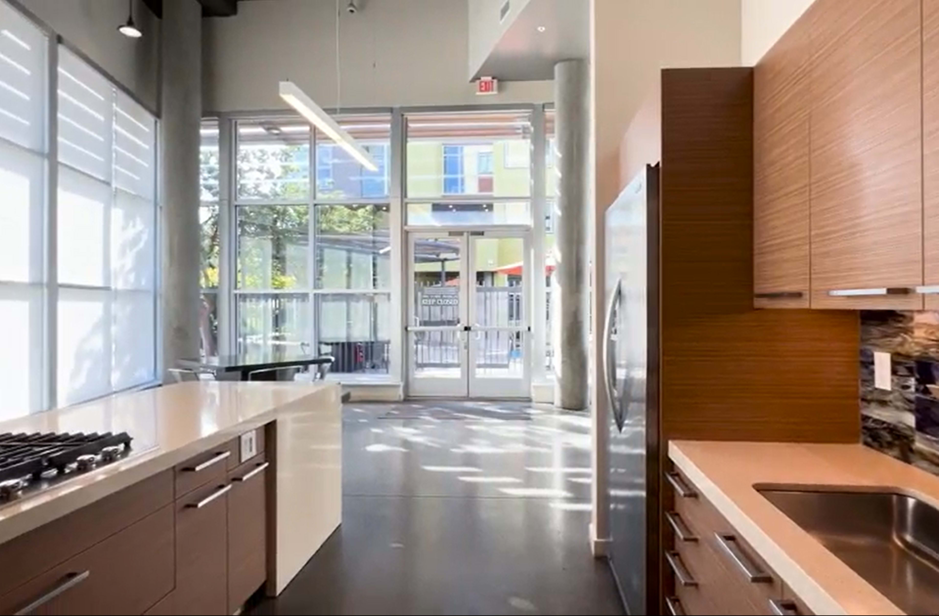 The image shows a modern kitchen with a long countertop, stainless steel appliances, and large windows allowing natural light to fill the space.