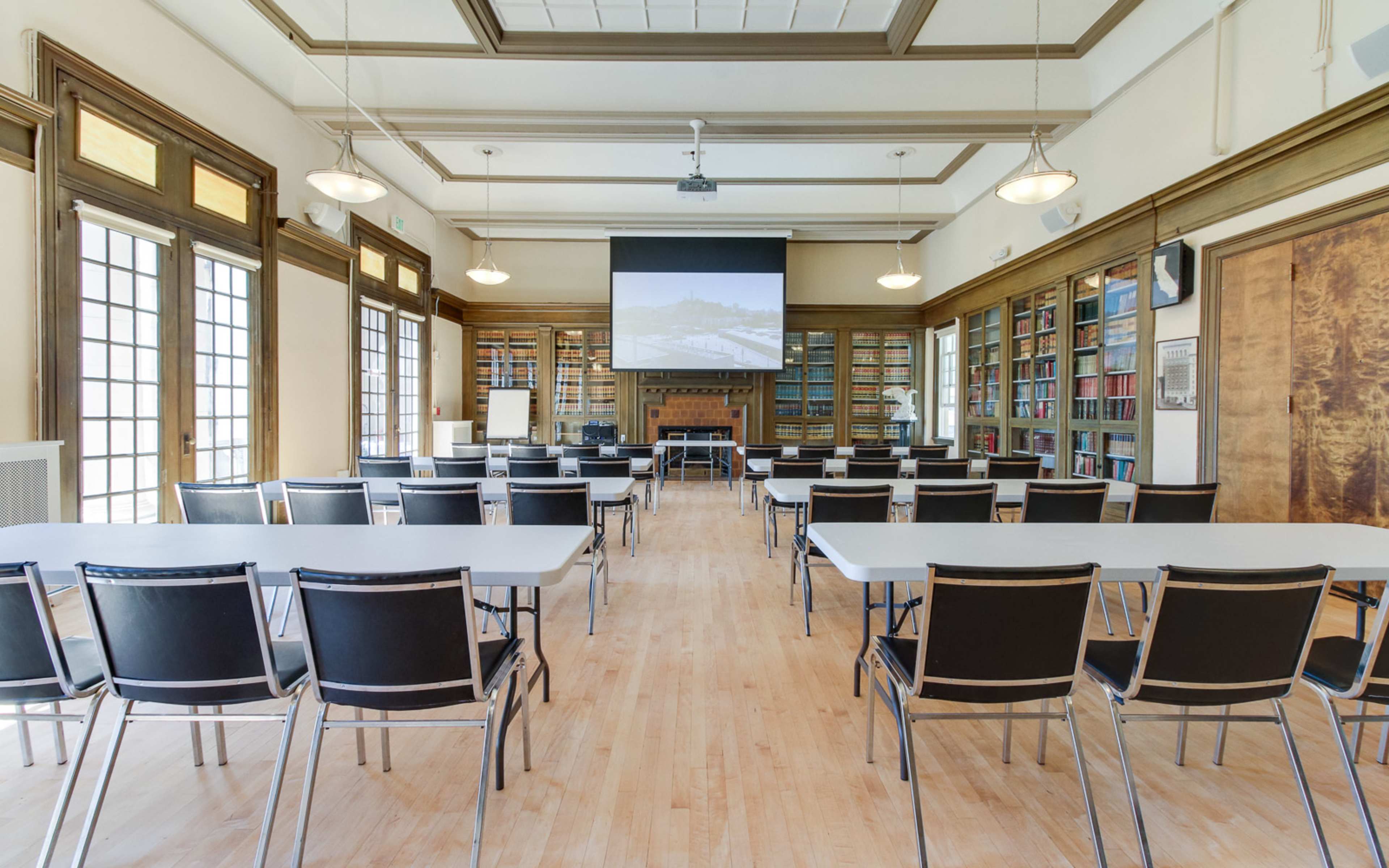 The image shows a spacious conference room with rows of black chairs facing a large projection screen, surrounded by bookshelves and windows.