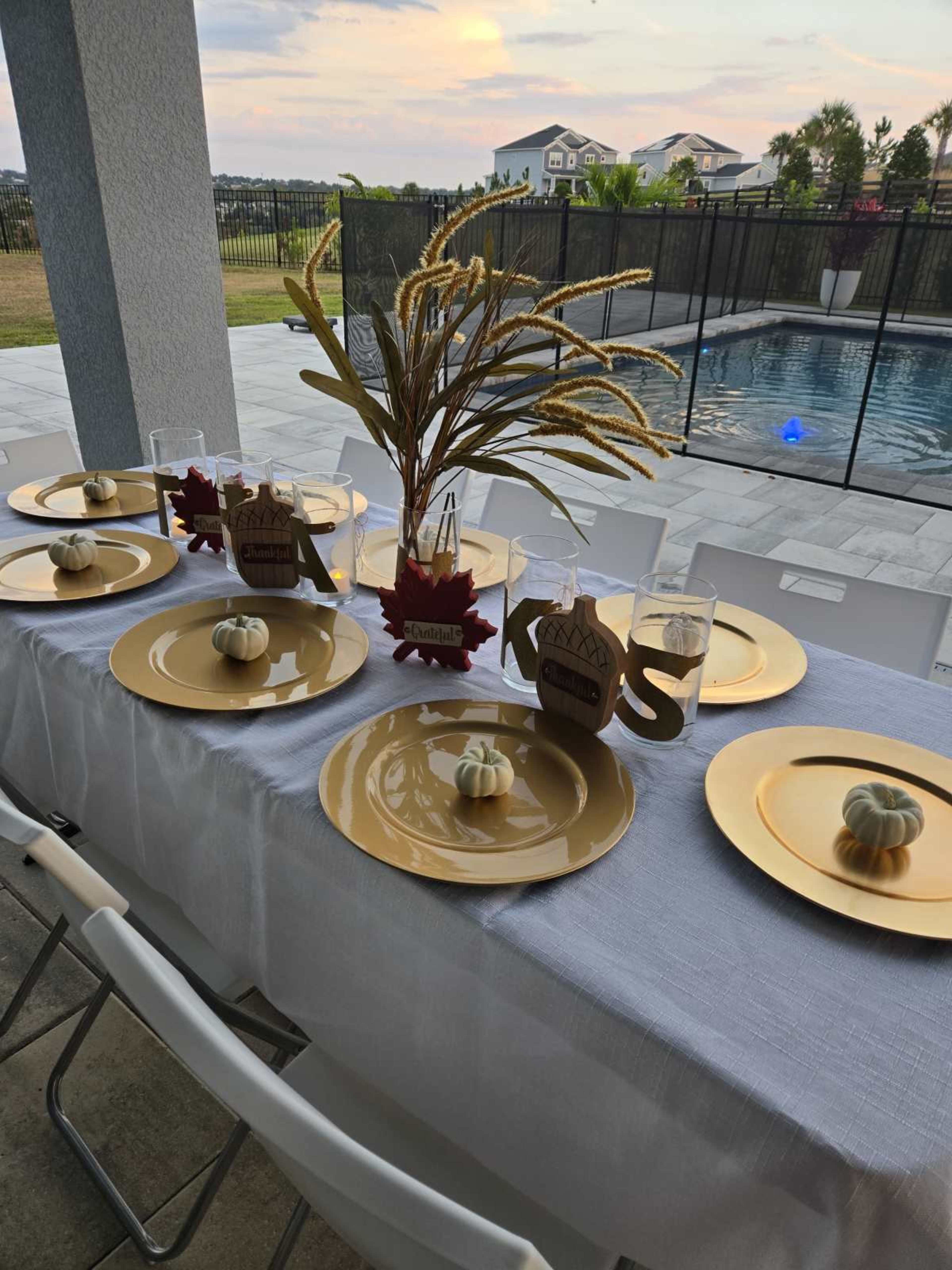 A dining table is set outdoors with gold plates, decorative pumpkins, and a centerpiece featuring dried wheat, overlooking a swimming pool.