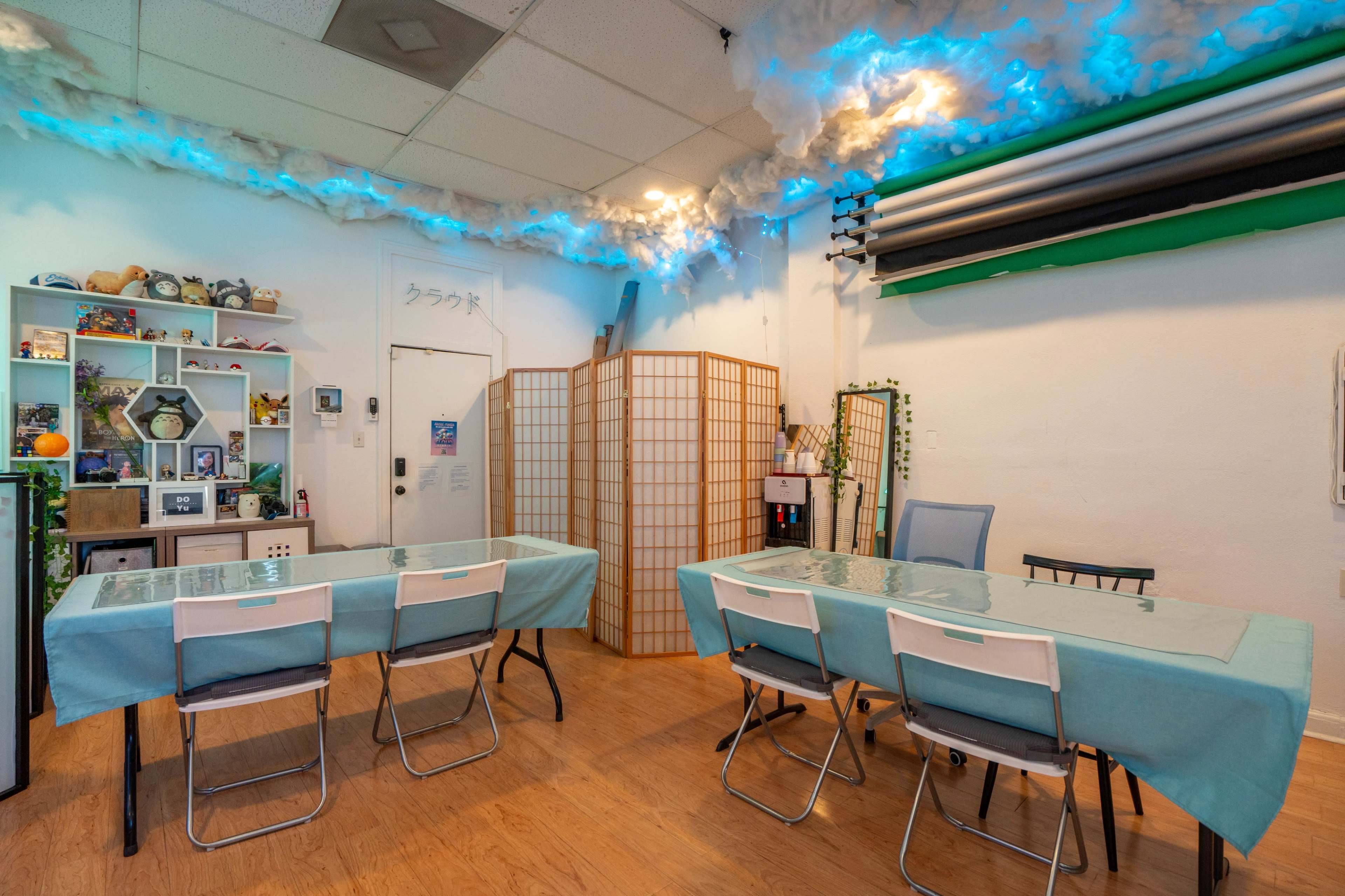 A brightly lit room features two tables covered in turquoise tablecloths, surrounded by chairs, with decorative shelves and partition screens in the background.
