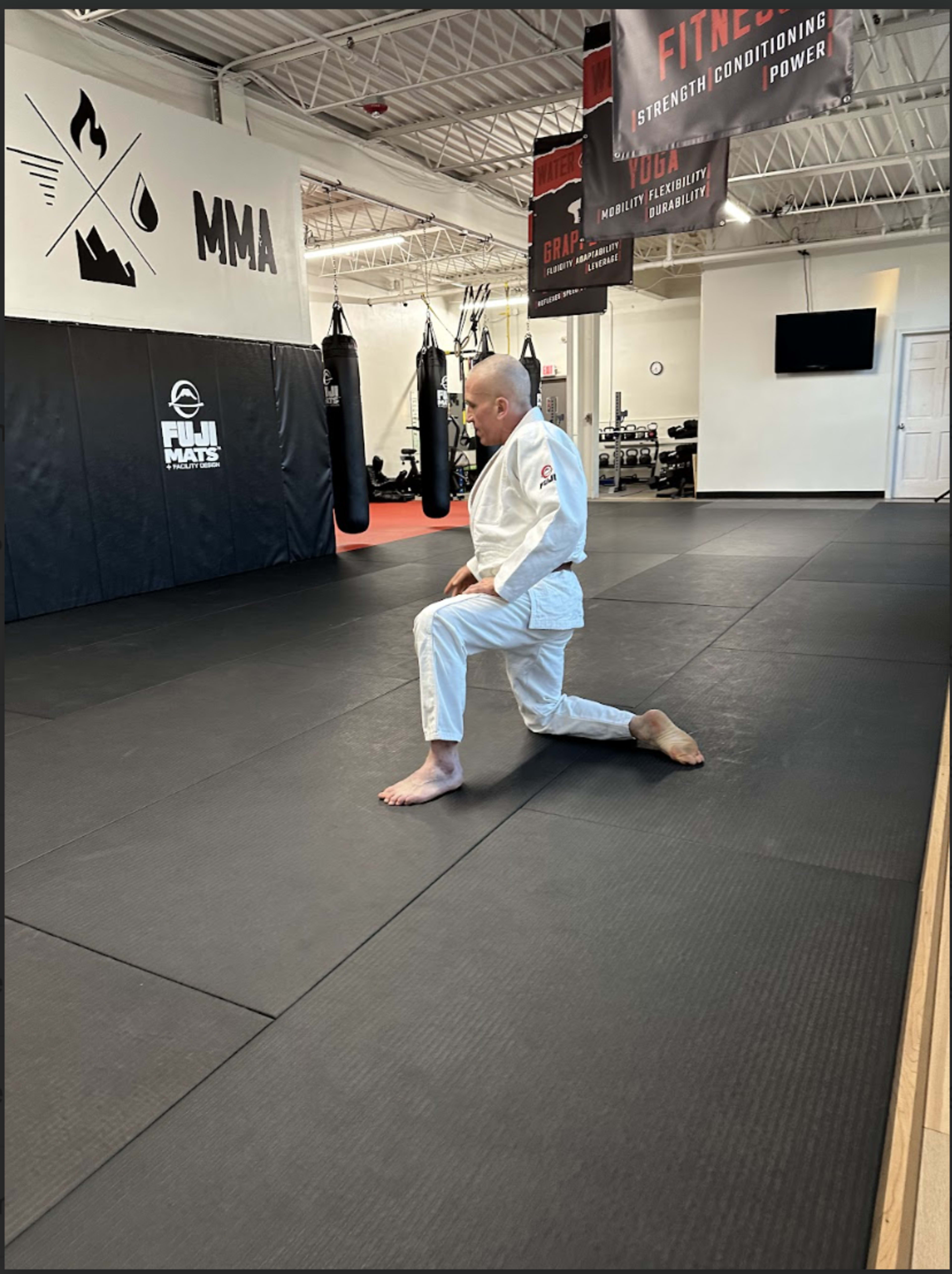 A person in a white jiu-jitsu uniform kneels on a black mat in a gym with workout equipment and punching bags in the background.