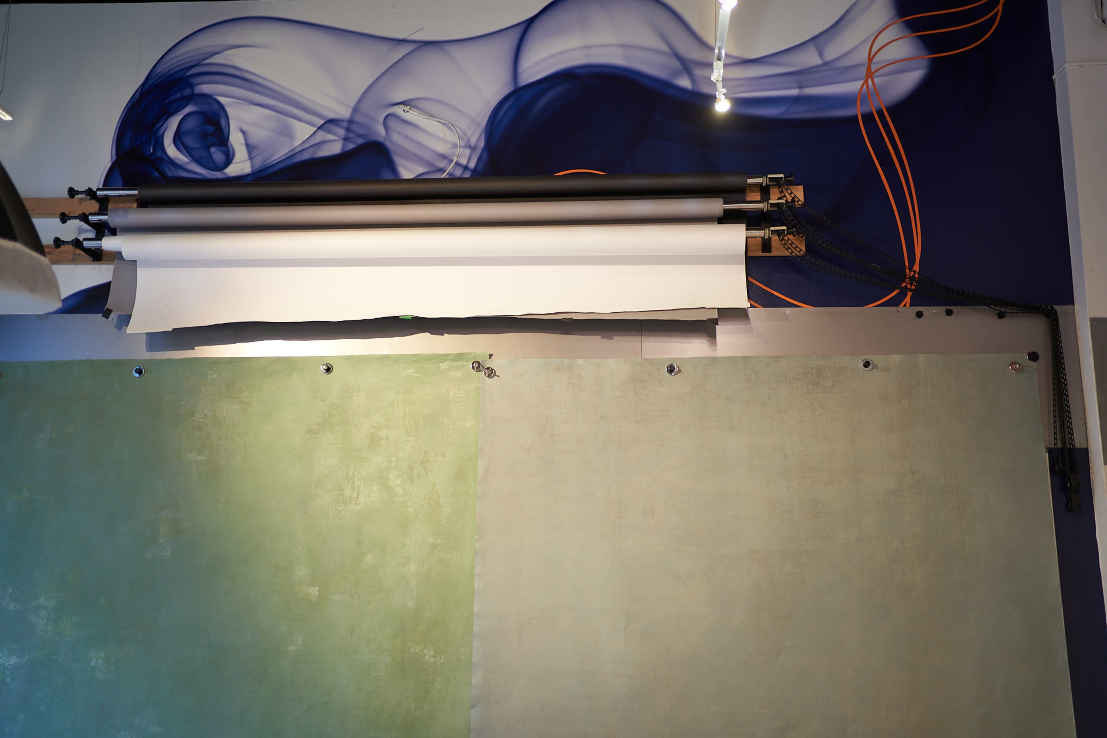 The image shows a studio backdrop setup with green and beige rolled paper mounted above a painted wall featuring a blue swirl design.