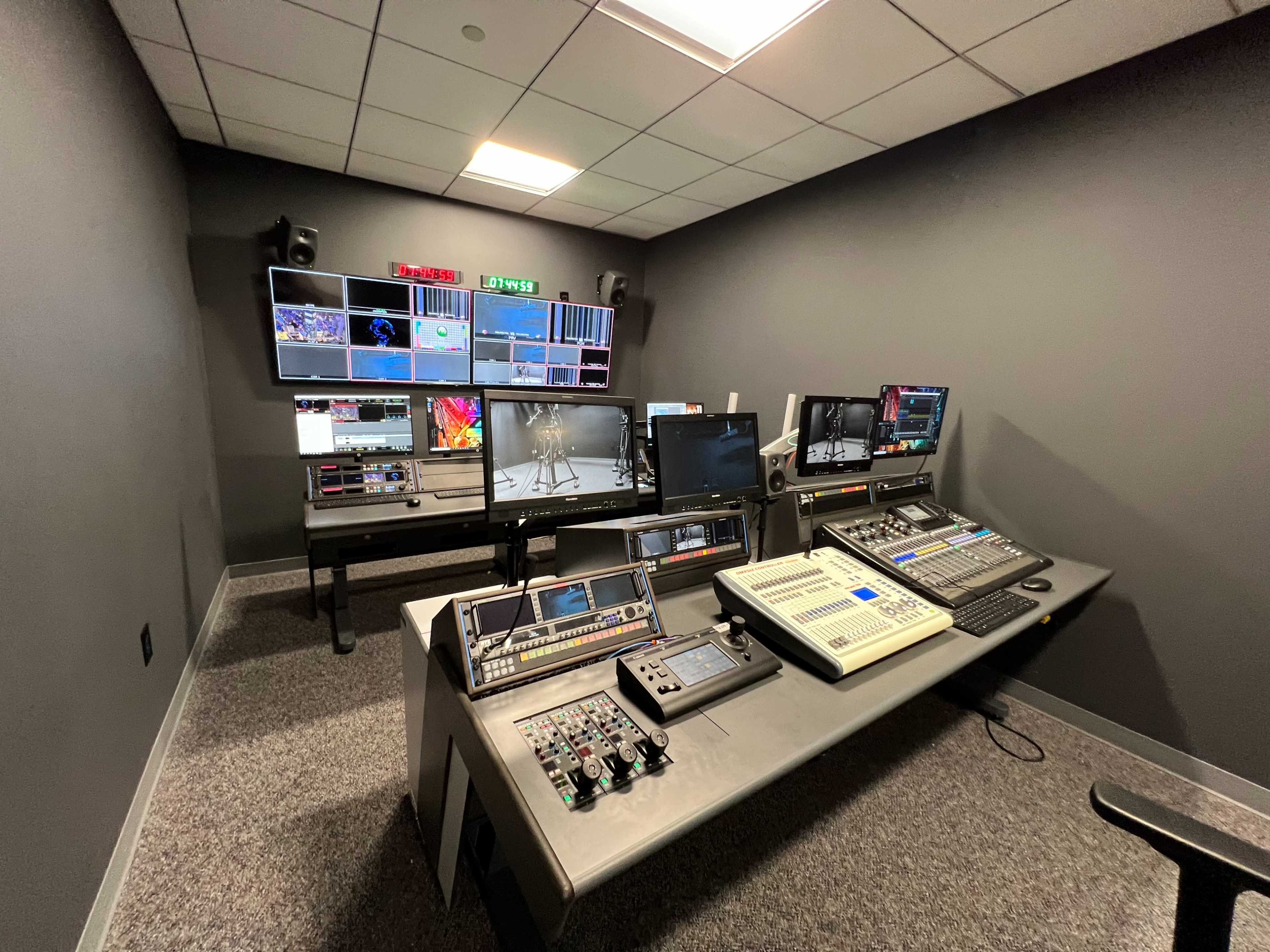 The image shows a broadcast control room equipped with multiple monitors, audio equipment, and a production console.