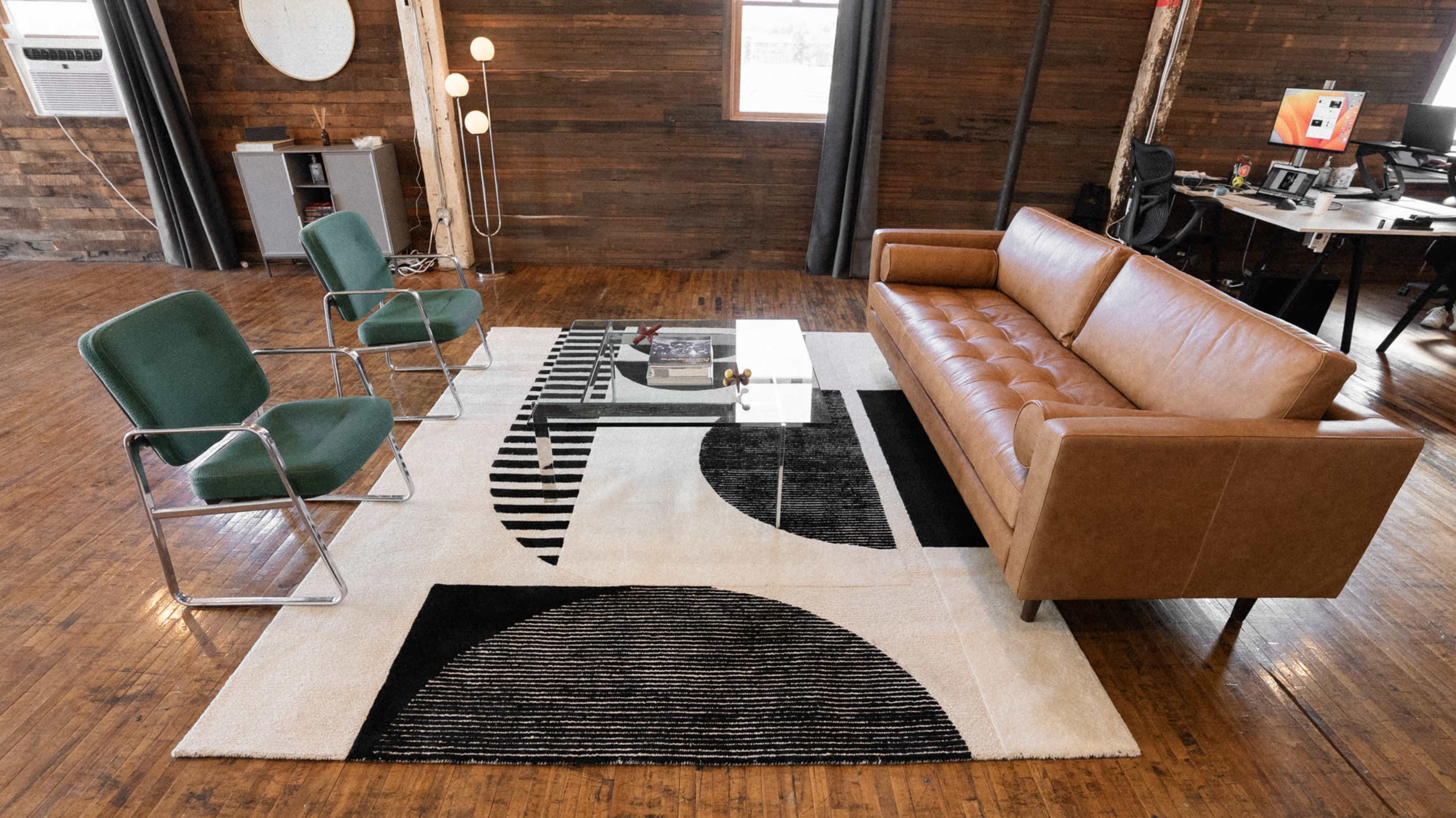 A modern living space features a brown leather sofa, green chairs, a glass coffee table, and a patterned rug, all set against a wooden wall.