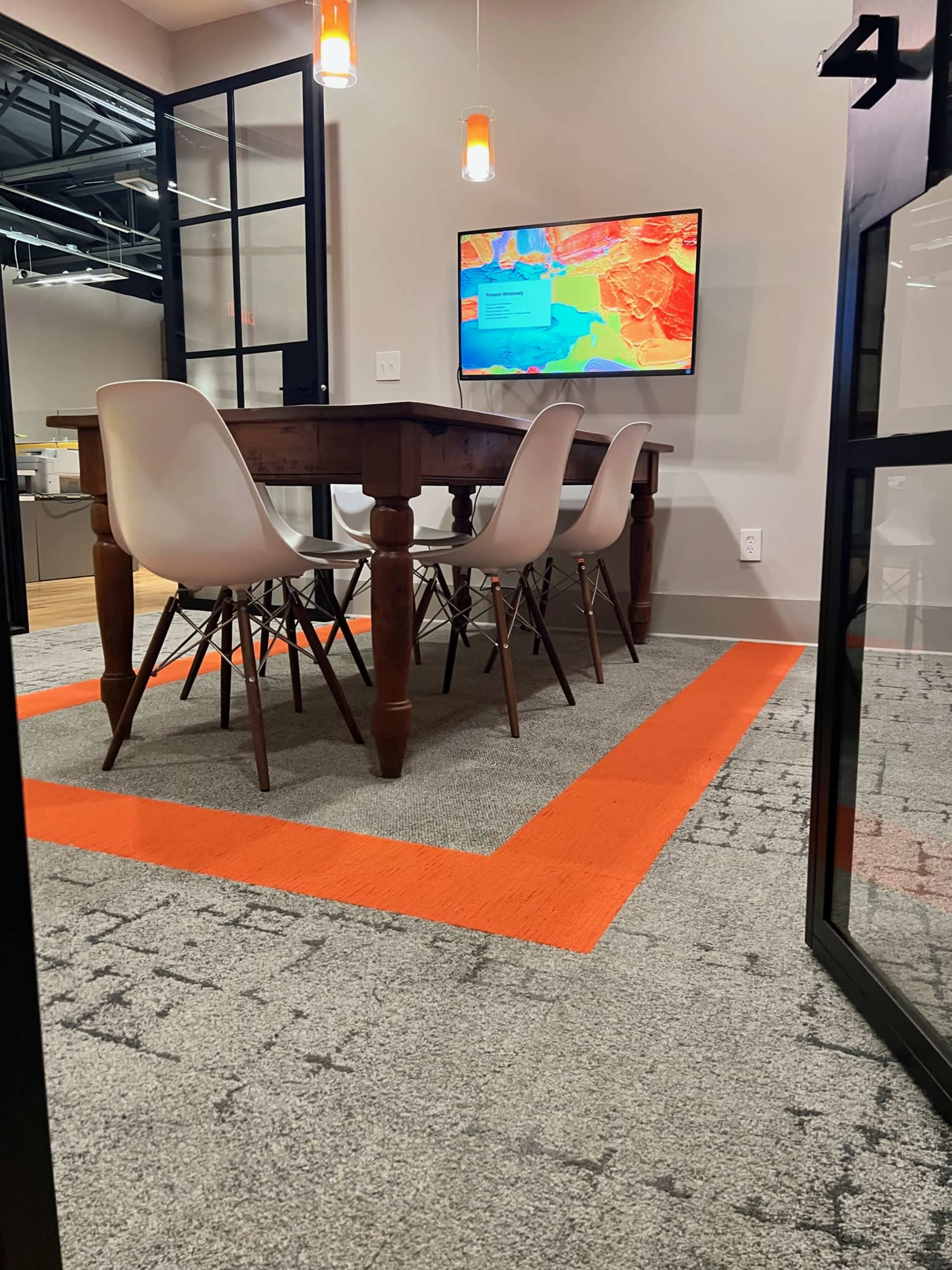 A wooden table surrounded by four white chairs sits in a meeting room with a television mounted on the wall displaying a colorful map.