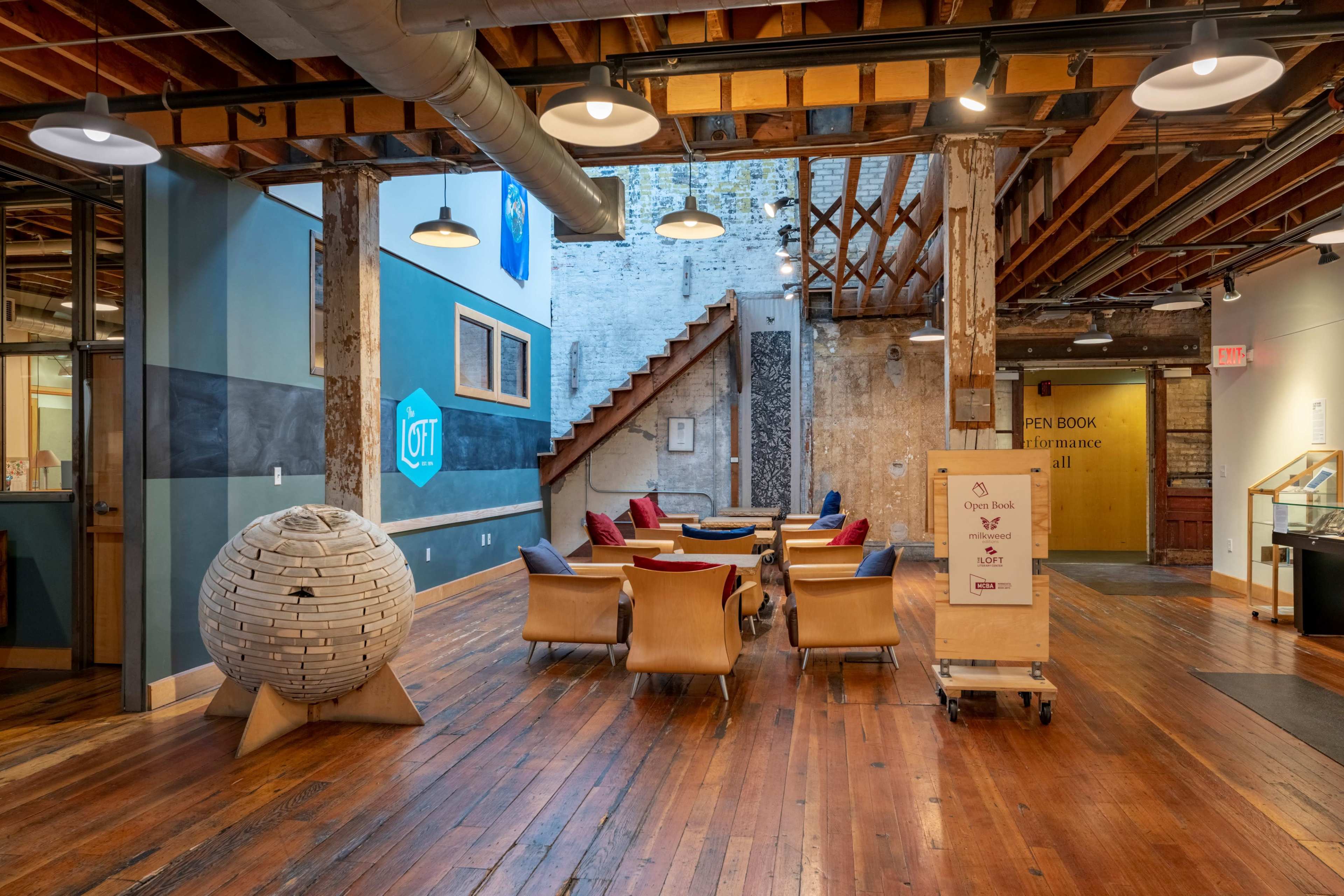 The image shows a spacious interior of a modern, open-concept area with wooden floors, a staircase in the background, and a seating arrangement featuring chairs and cushions.