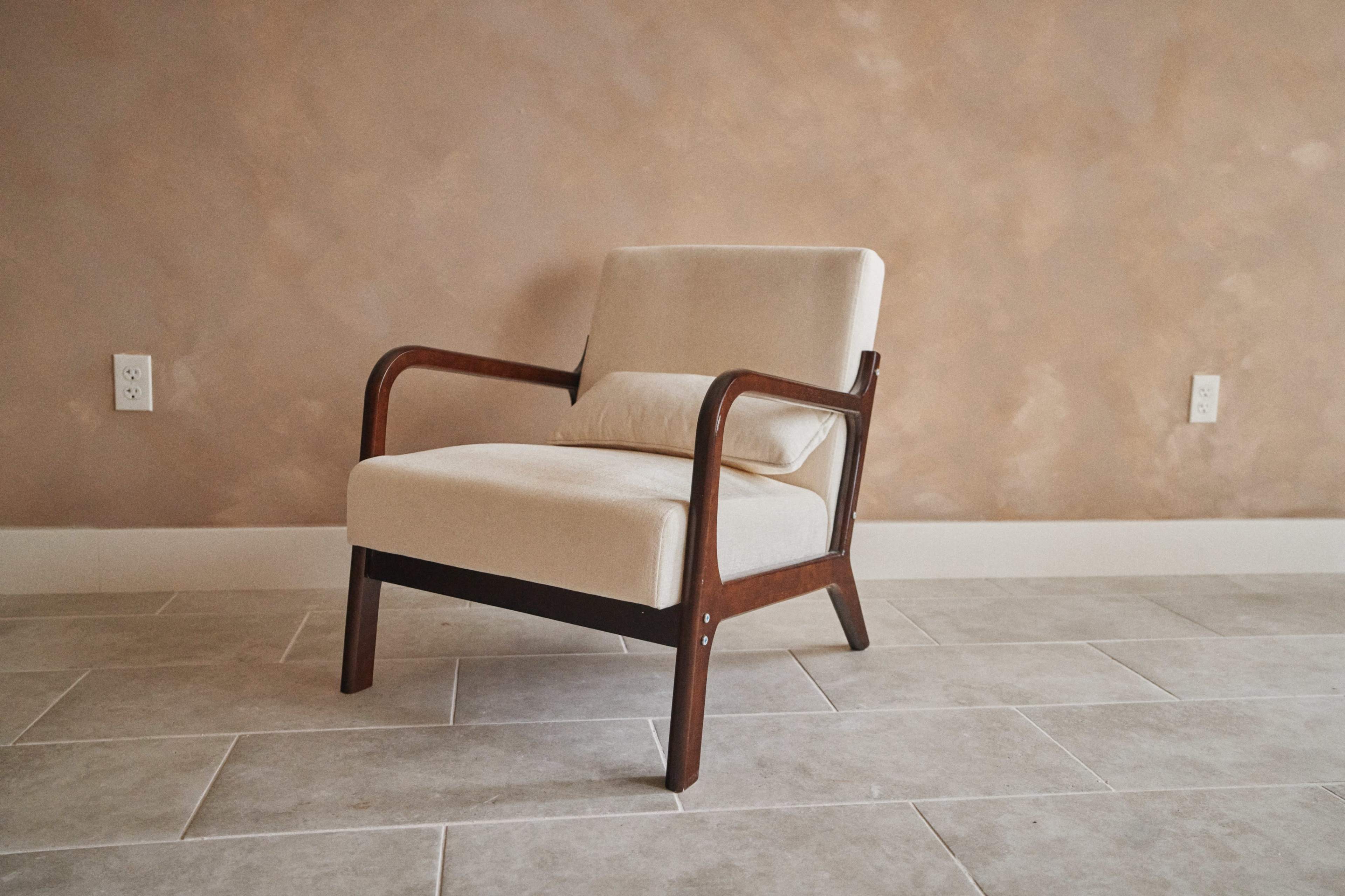 A beige upholstered chair with wooden armrests sits on a light-colored tiled floor against a textured, warm-toned wall.