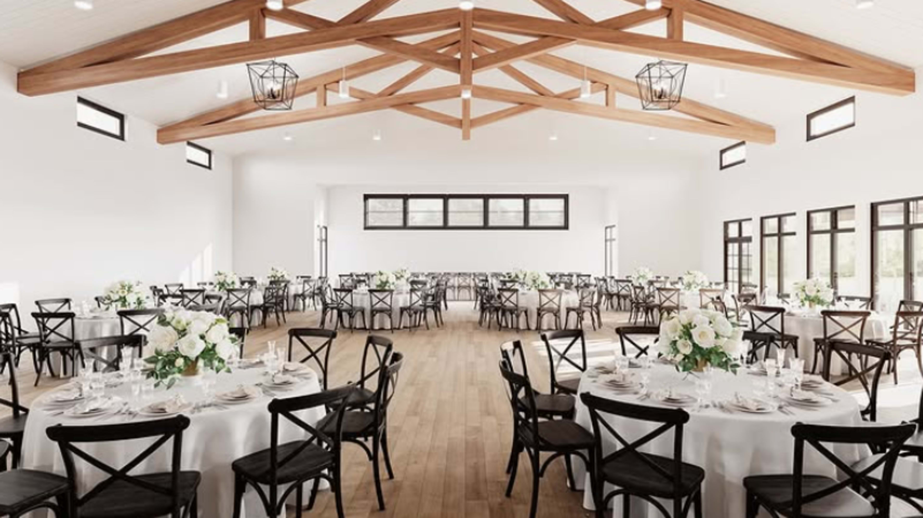 The image shows a spacious banquet hall set with round tables and floral centerpieces, featuring a wooden beam ceiling and large windows.
