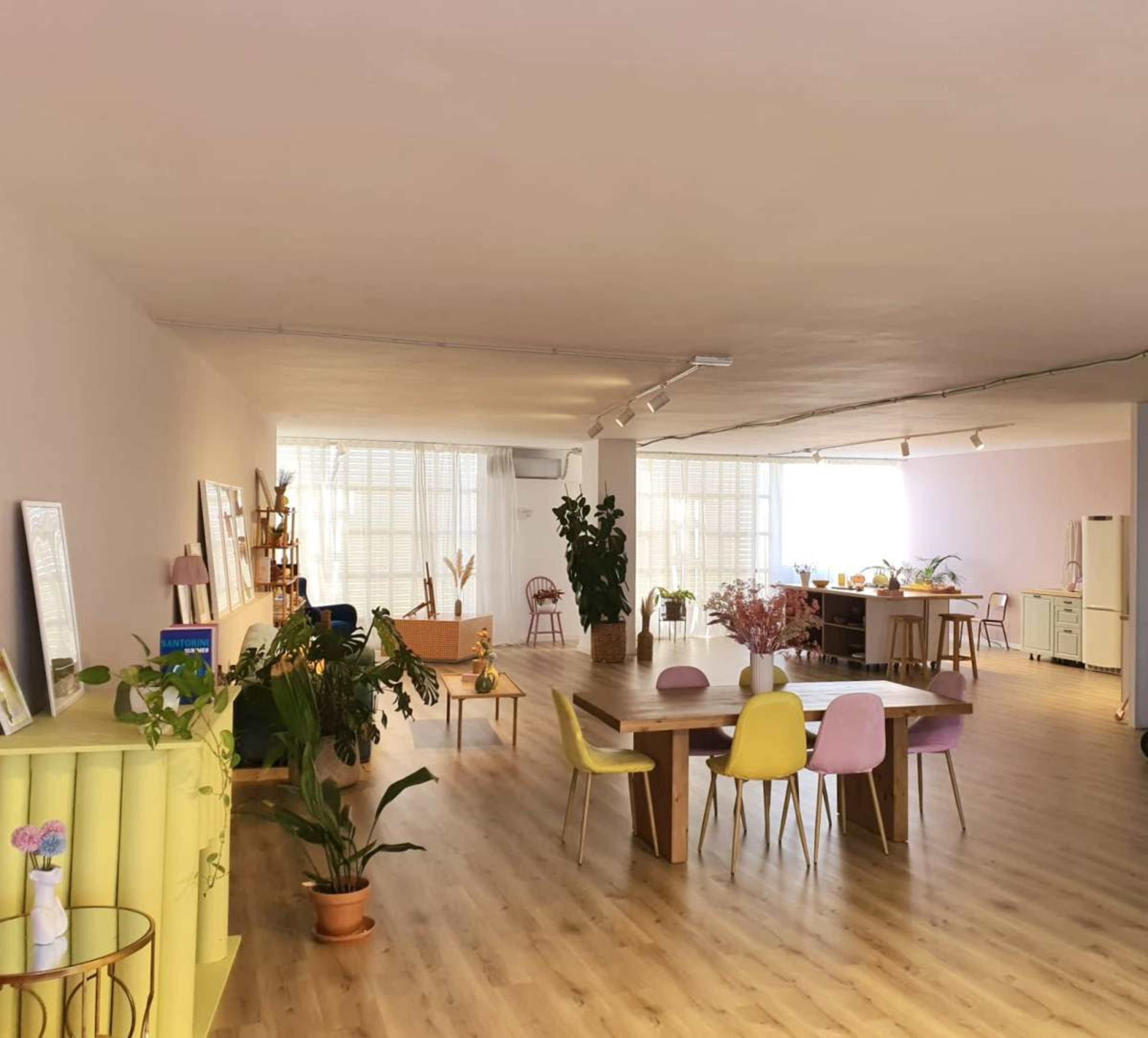 A spacious, bright room features a large wooden table surrounded by colorful chairs, plants, and various workstations.